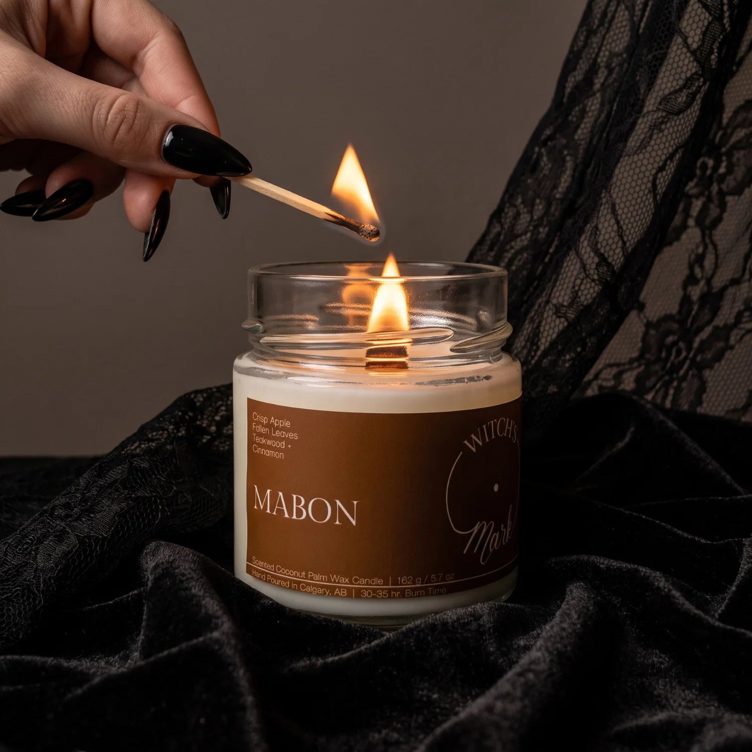 A woman's hand with black fingernails lights a candle called "Mabon" with a match. The candle rests on a bed of black lace fabric.