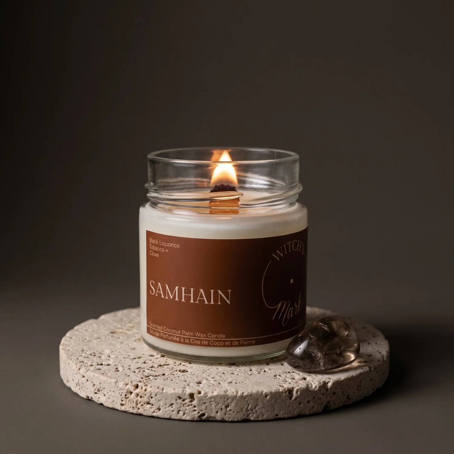A Witch's Mark Samhain crystal candle sits on top of a porous stone coaster with a smoky quartz crystal beside it.