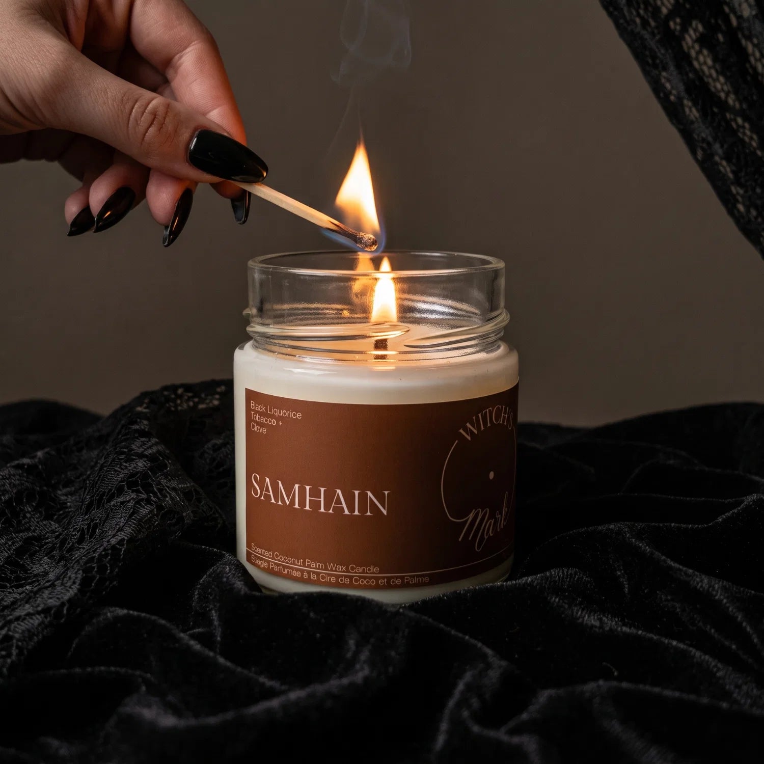 A woman's hand with black fingernails lights a candle called "Samhain" with a match. The candle sits on black lace fabric that draped around.