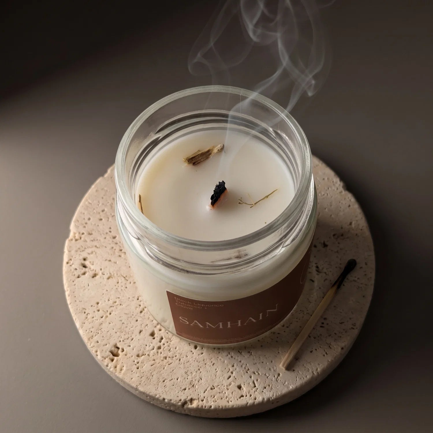 A Samhain candle sits on a porous stone coaster with it's wick recently extinguished and curls of white smoke rising.
