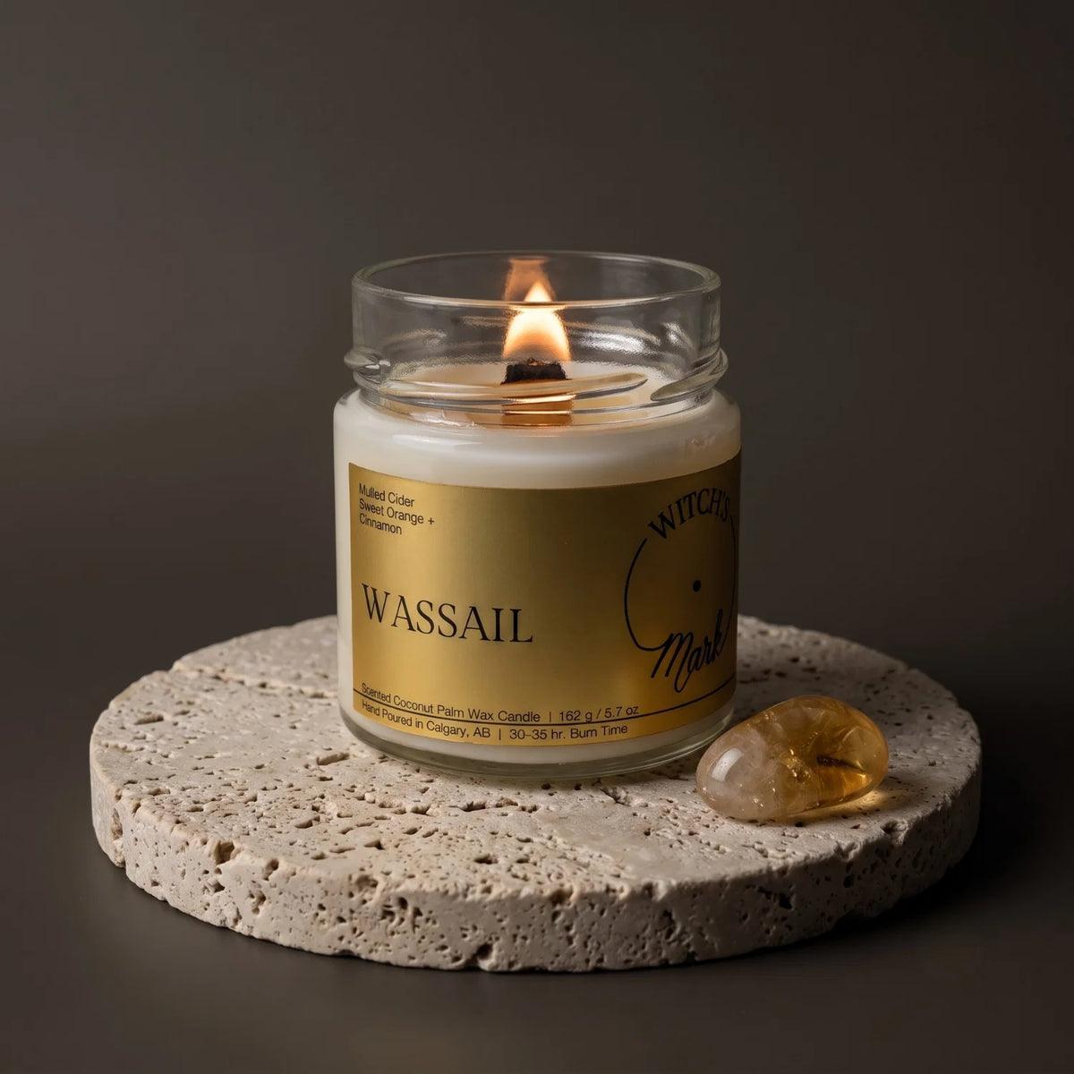 A Witch's Mark candle called "Wassail" with a gold label sits lit on a porous stone coaster with a citrine crystal beside it.