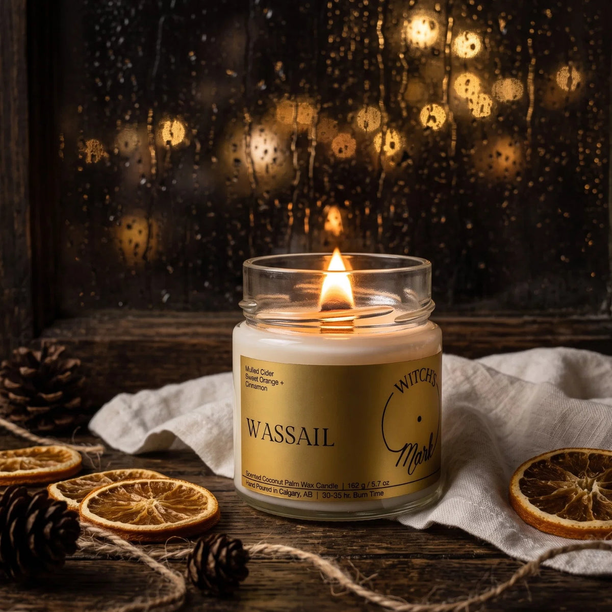 A lit Witch's Mark Wassail candle sits on a rustic wooden surface with dried orange slices, pinecones, and a white cloth. The background shows a dark rainy window with warm golden bokeh lights.