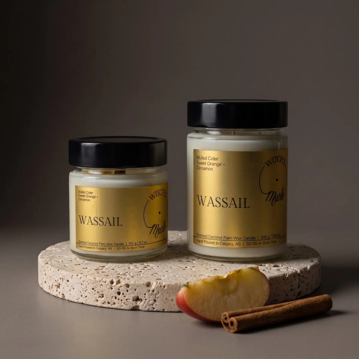Two Wassail scented candles in clear glass jars with black lids and gold labels, displayed on a round stone slab with a sliced apple and cinnamon stick against a dark gray background.