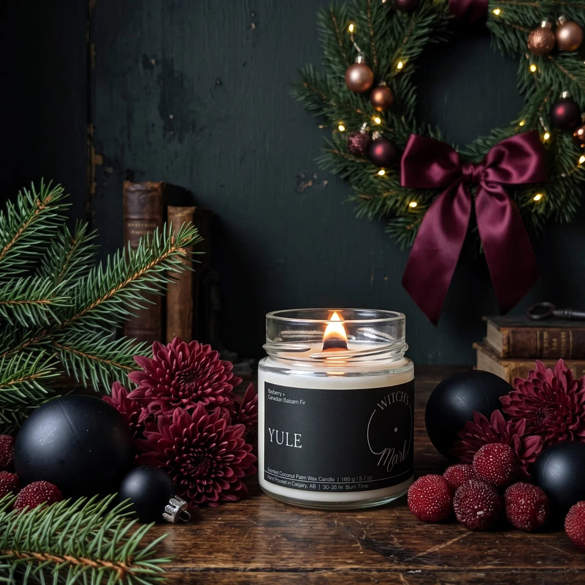 A lit Yule candle in a clear glass jar sits on a rustic wooden table surrounded by Christmas greenery, black ornaments, burgundy flowers, and a dark green wreath with a large burgundy bow in the background.