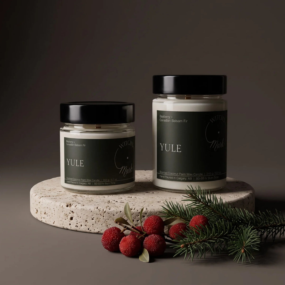 Two Yule candles sit on a porous stone coaster with bayberries and a balsam branch in front of the coaster.