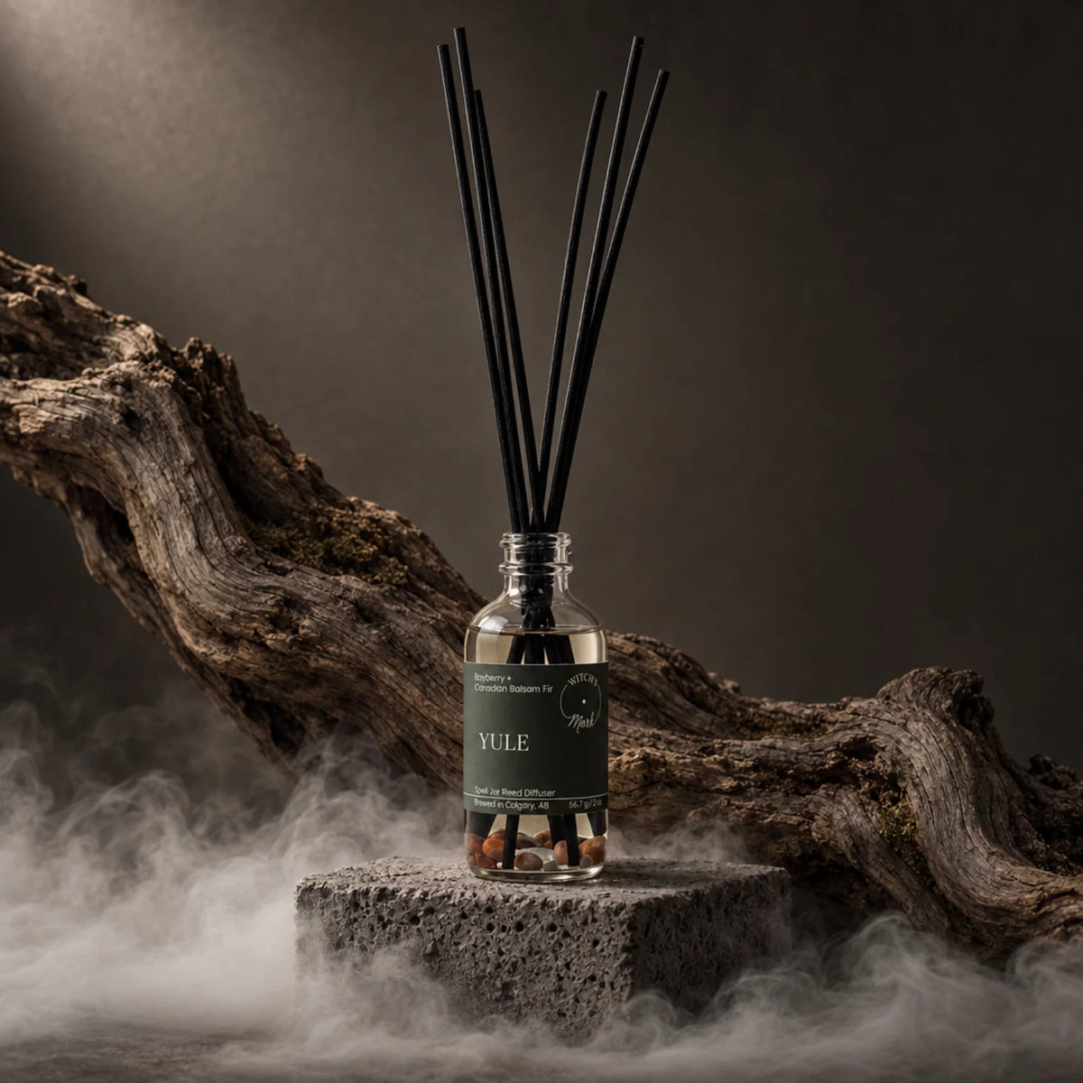 A reed diffuser bottle with a dark green label reading YULE, filled with clear liquid and black reeds, displayed on a stone pedestal with a large piece of driftwood and mist in the background.