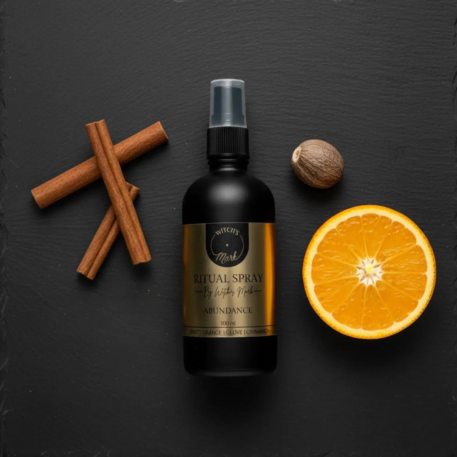 A top-down flat lay highlighting the warm, wealthy ingredients of the Abundance ritual mist by Witch's Mark. The black apothecary bottle with a gold label sits surrounded by spiced cinnamon sticks, whole cloves, and a slice of bright sweet orange against a dark slate background.