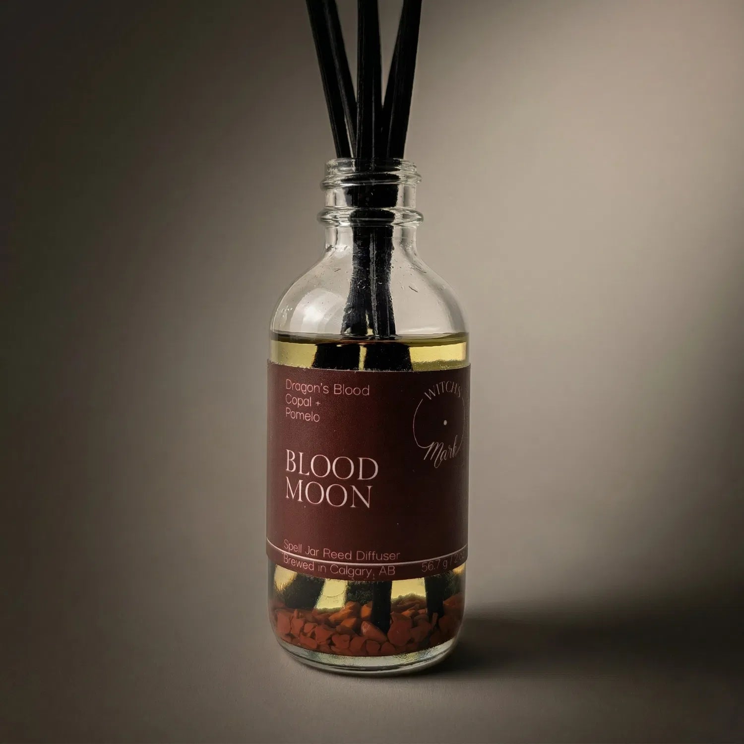 A dramatic spotlight close-up of the Blood Moon spell jar reed diffuser, highlighting the deep red front label which lists its magical, fortifying scent profile of ancient dragon's blood, sacred copal smoke, and bright pomelo.