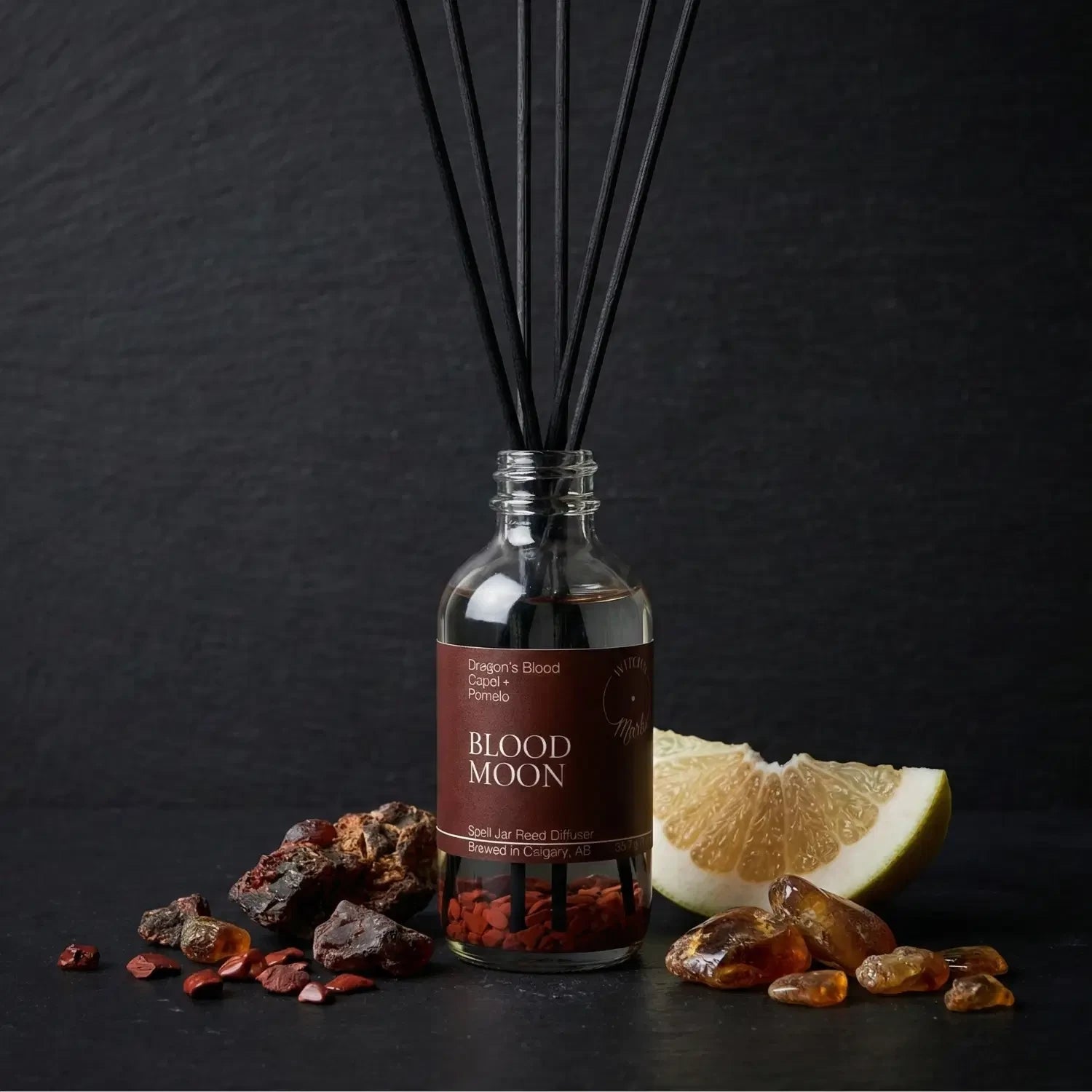 The Blood Moon spell jar reed diffuser by Witch's Mark showcased against a dark slate background, surrounded by its core empowering botanical ingredients: dark red dragon's blood resin, golden chunks of sacred copal, and a slice of bright pomelo citrus.