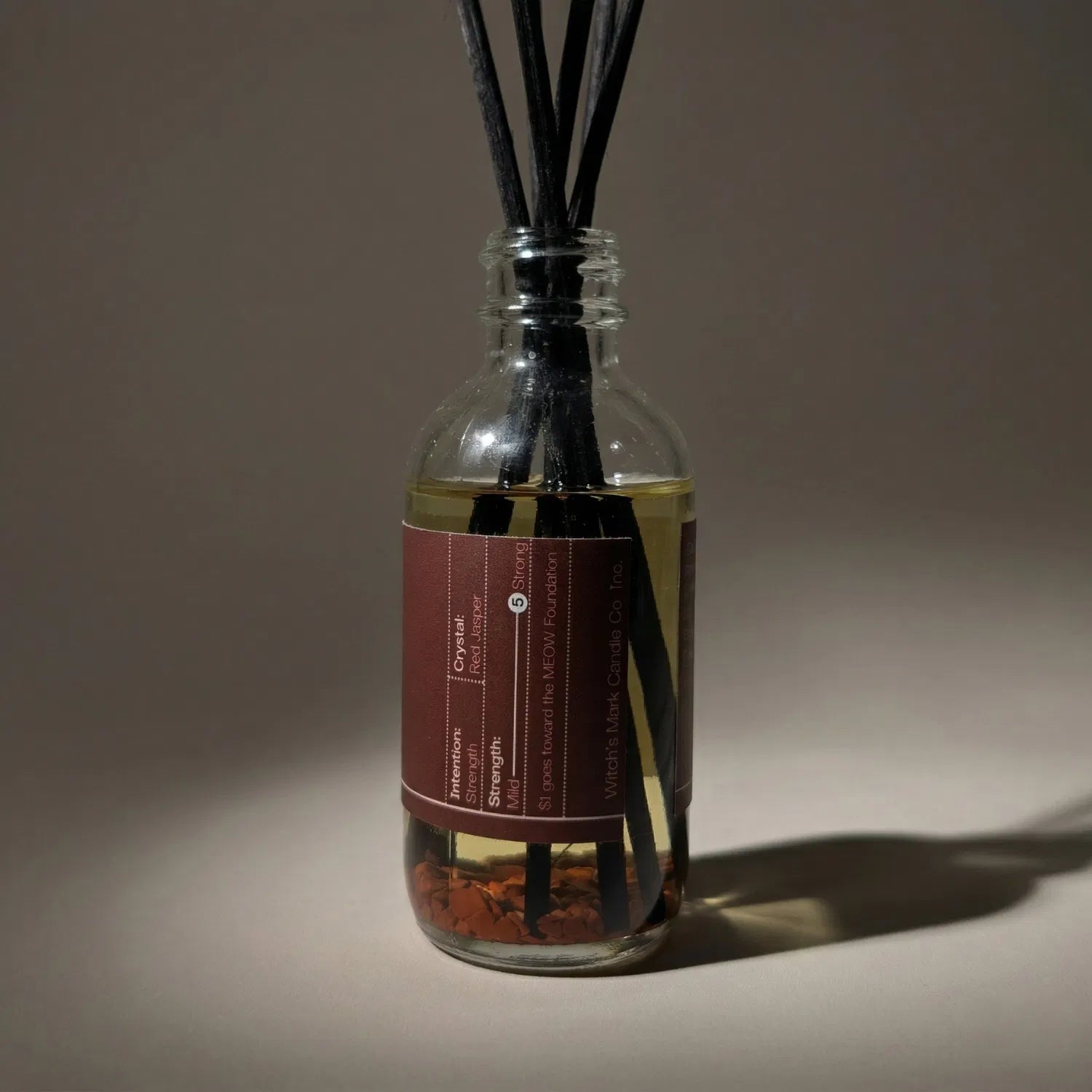 A warmly lit close-up of the Blood Moon spell jar reed diffuser, revealing the fierce red jasper crystals submerged at the bottom of the golden botanical oil, crafted to invoke supreme endurance and deeply rooted strength.