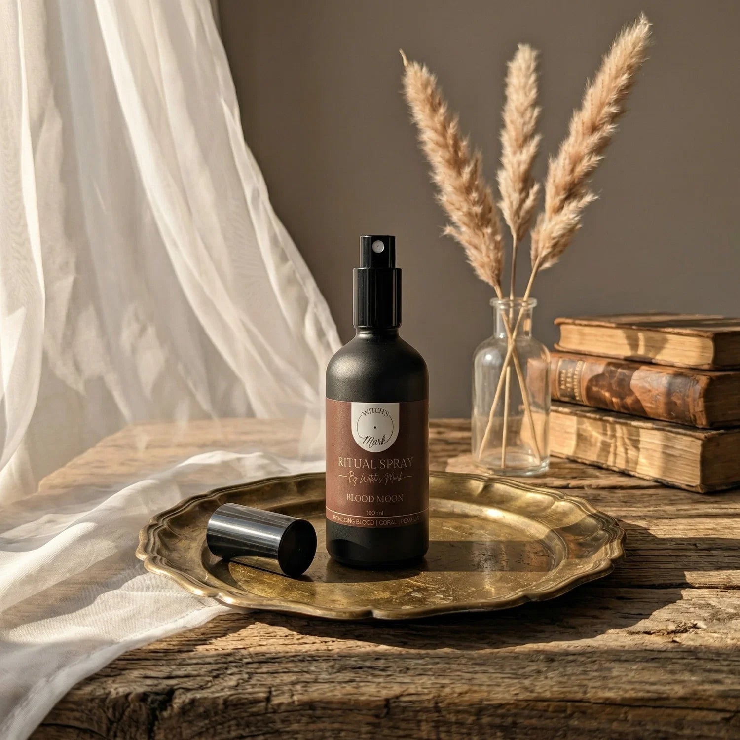 The matte black bottle of Blood Moon ritual spray with its deep red label elegantly displayed on a vintage brass tray atop a rustic wooden table bathed in soft light. This sacred elixir is perfect for misting over your altar to anchor your boldest spellwork and protection rites.