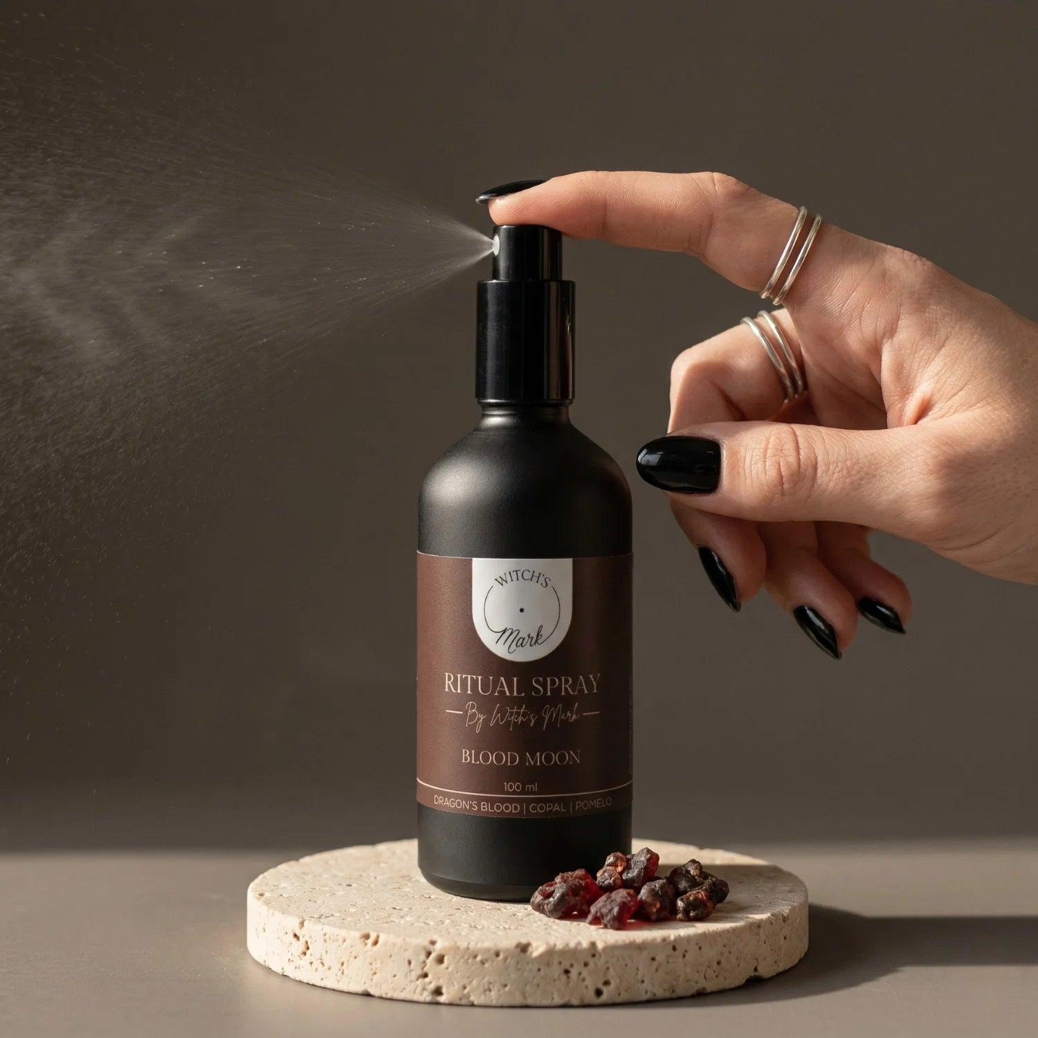 A hand with black nail polish spritzing the Witch's Mark Blood Moon ritual spray over scattered chunks of dark red dragon's blood resin. This potent aura mist is infused with red jasper charged moon water and rich resins to awaken elemental fire and fortify the spirit.