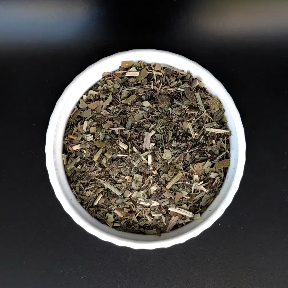 A top-down, close-up view of the Clairvoy-Mint loose leaf herbal tea blend by Witch's Mark in a small white bowl. The image clearly showcases the high-quality mix of dried yerba mate, cut lemongrass, ginkgo, and peppermint against a dark background.