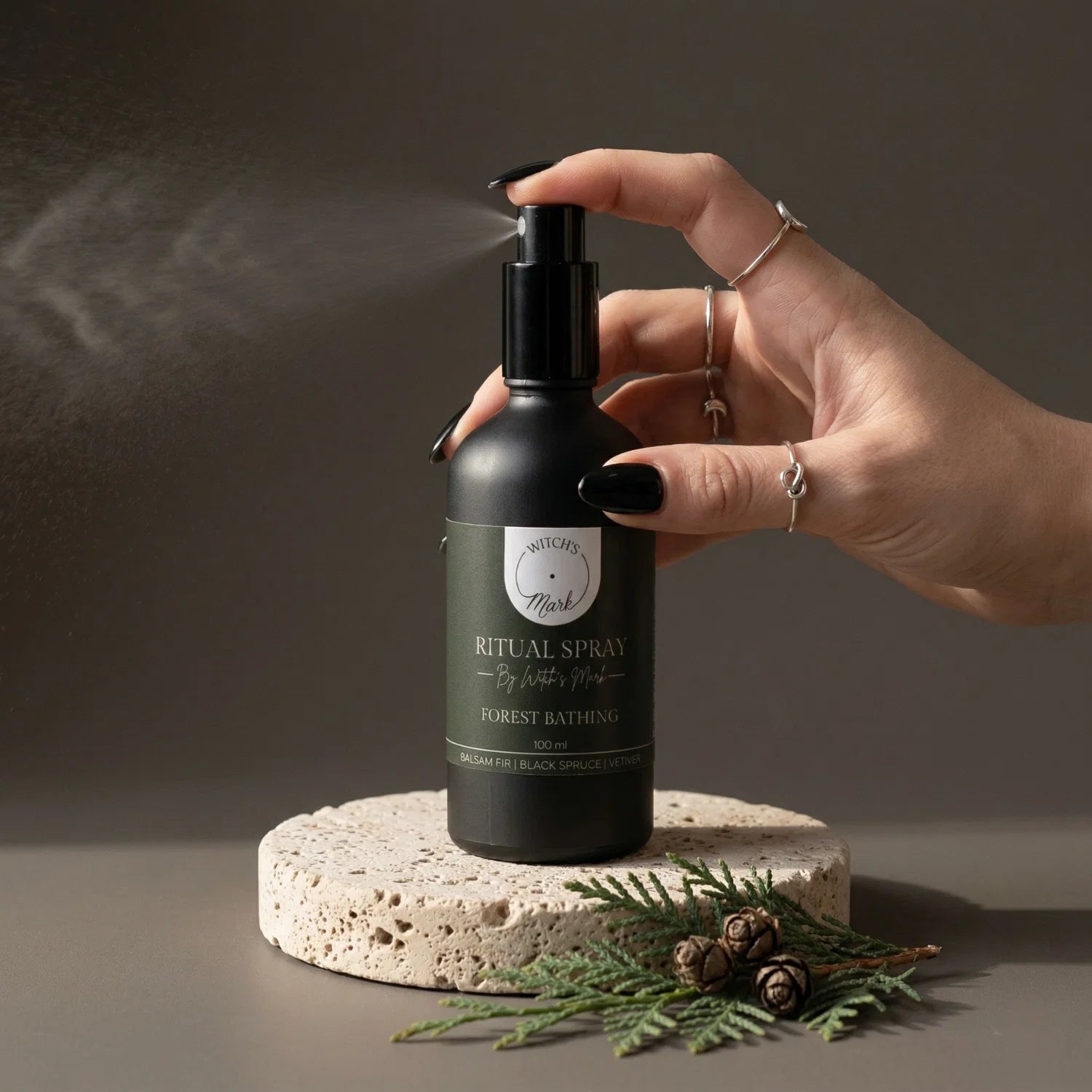 A hand with black nail polish spritzing the Forest Bathing ritual spray by Witch's Mark to cleanse the aura and room, set next to a natural pinecone and fresh spruce sprig.
