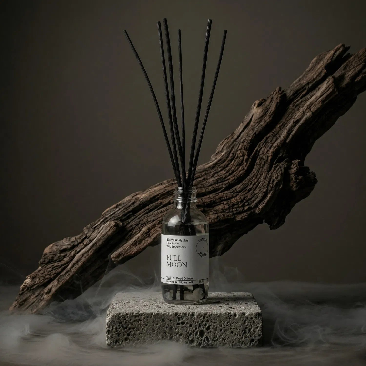 The Full Moon apothecary spell jar reed diffuser by Witch's Mark sitting on a stone base, enveloped in atmospheric mist with natural driftwood in the background, perfectly setting the stage for peak spiritual power and raw manifesting energy.