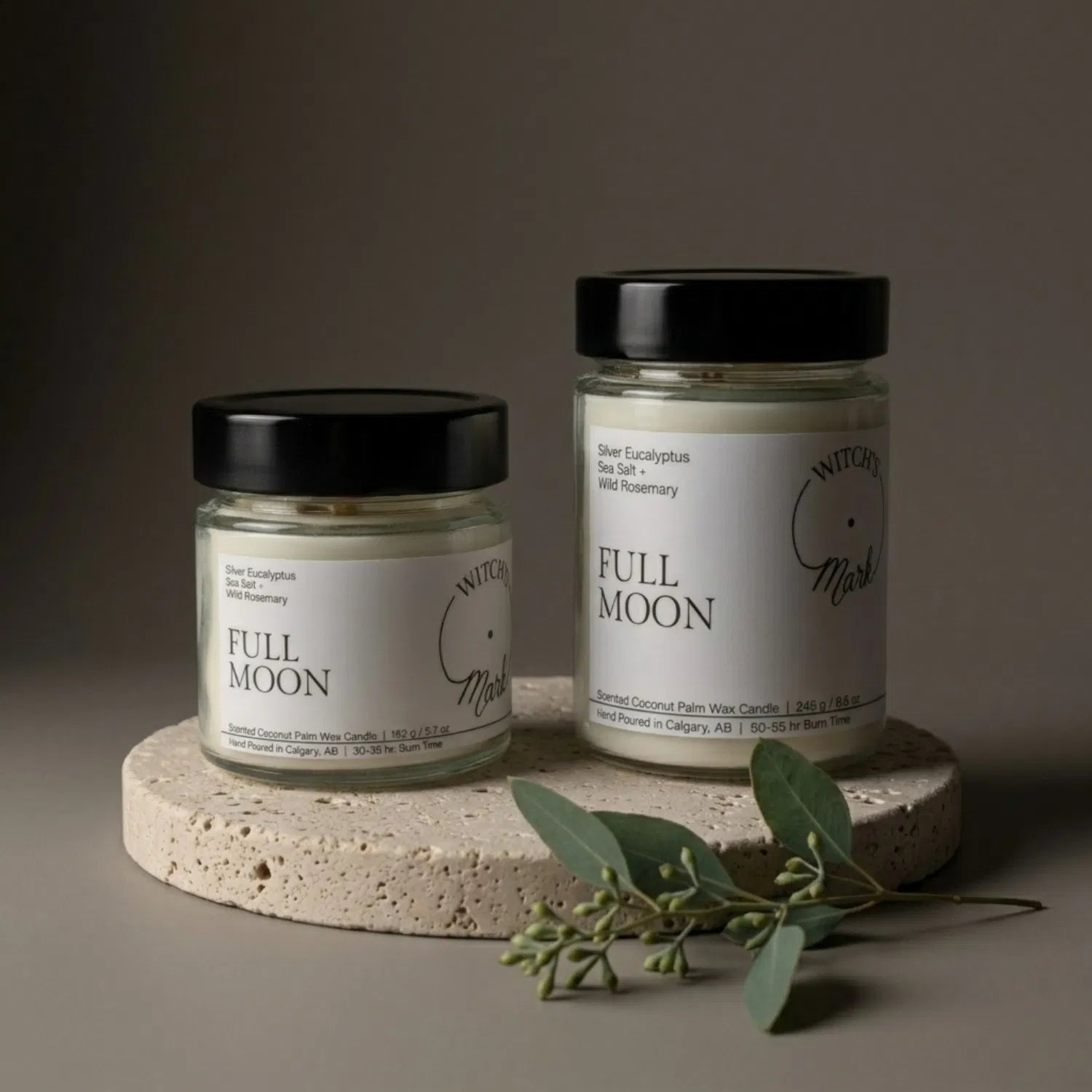 Two sizes of the Witch's Mark Full Moon coconut wax candle, 5.7 oz and 8.6 oz, displayed on a round stone base and garnished with fresh silver eucalyptus leaves.