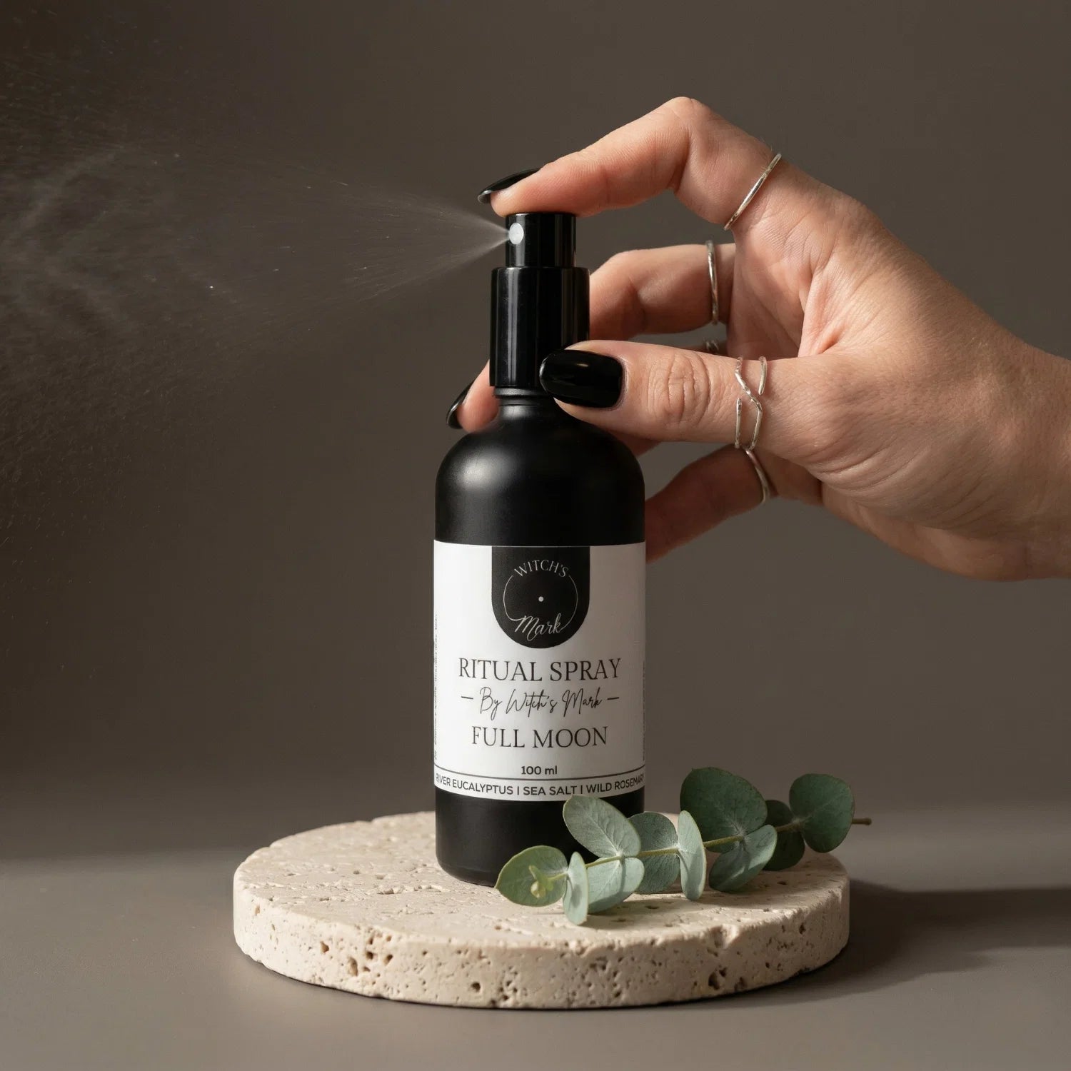 A hand with black nail polish spritzing the Witch's Mark Full Moon ritual spray over a fresh sprig of silver eucalyptus. This amplifying aura mist is infused with moonstone-charged moon water to awaken vibrant awareness and capture the raw energy of ultimate manifestation.
