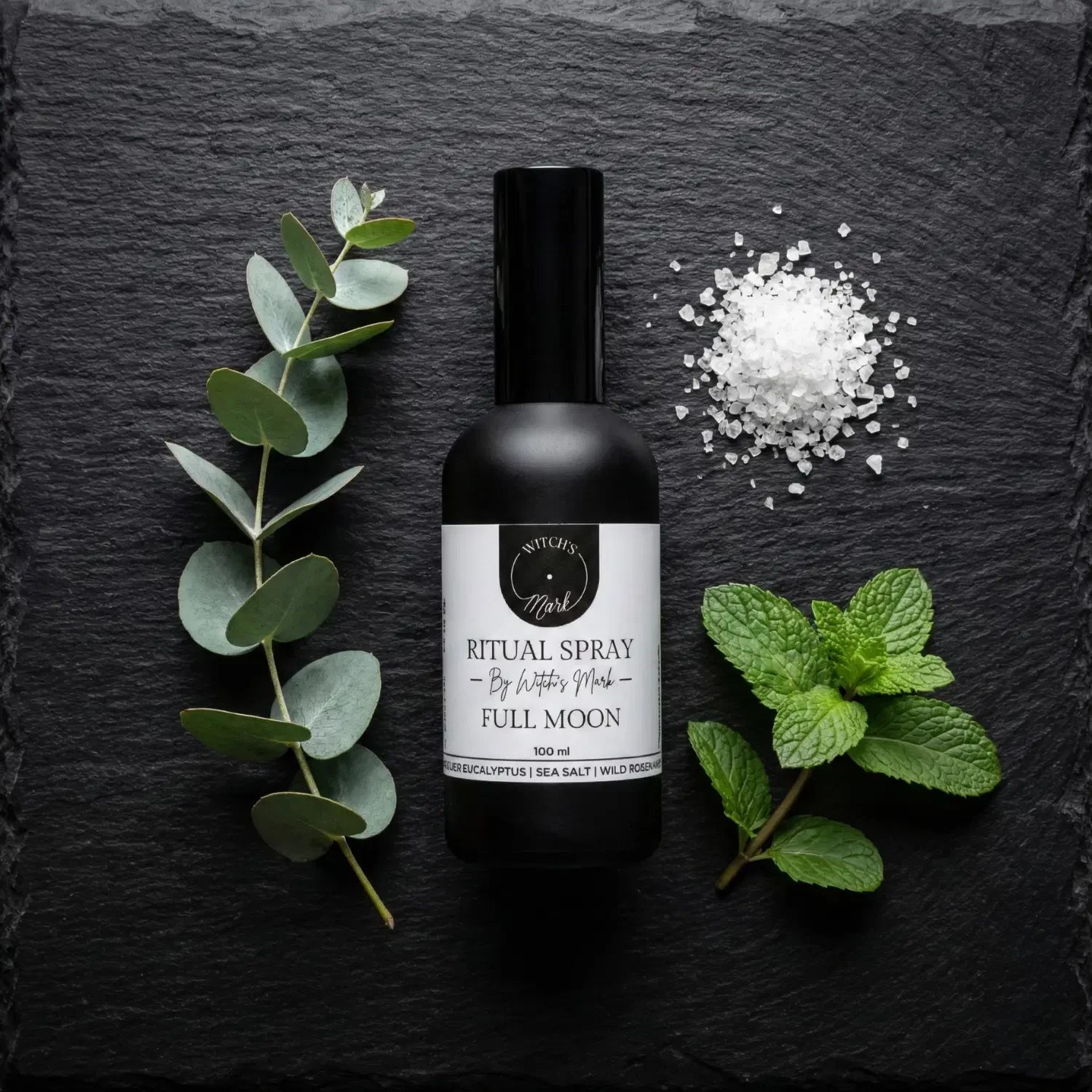A top-down flat lay highlighting the purifying ingredients of the Full Moon ritual mist by Witch's Mark. The black apothecary bottle sits surrounded by a fresh silver eucalyptus sprig, a pile of coarse sea salt, and bright green wild rosemary against a dark slate background.