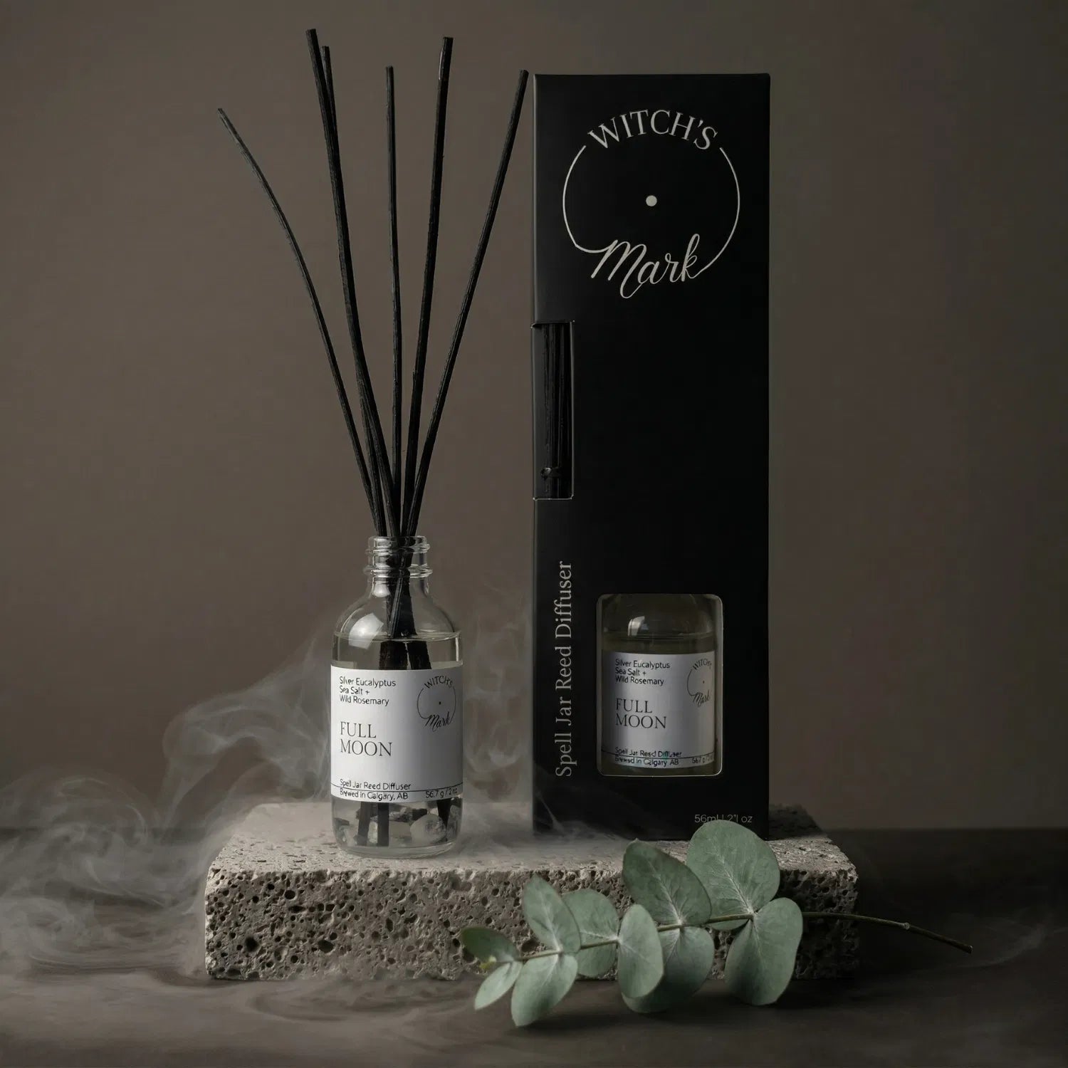 The Full Moon spell jar reed diffuser by Witch's Mark displayed next to its sleek black packaging box. The apothecary glass vessel with black reeds rests on a textured stone block surrounded by mystical mist and a fresh sprig of clarifying silver eucalyptus.
