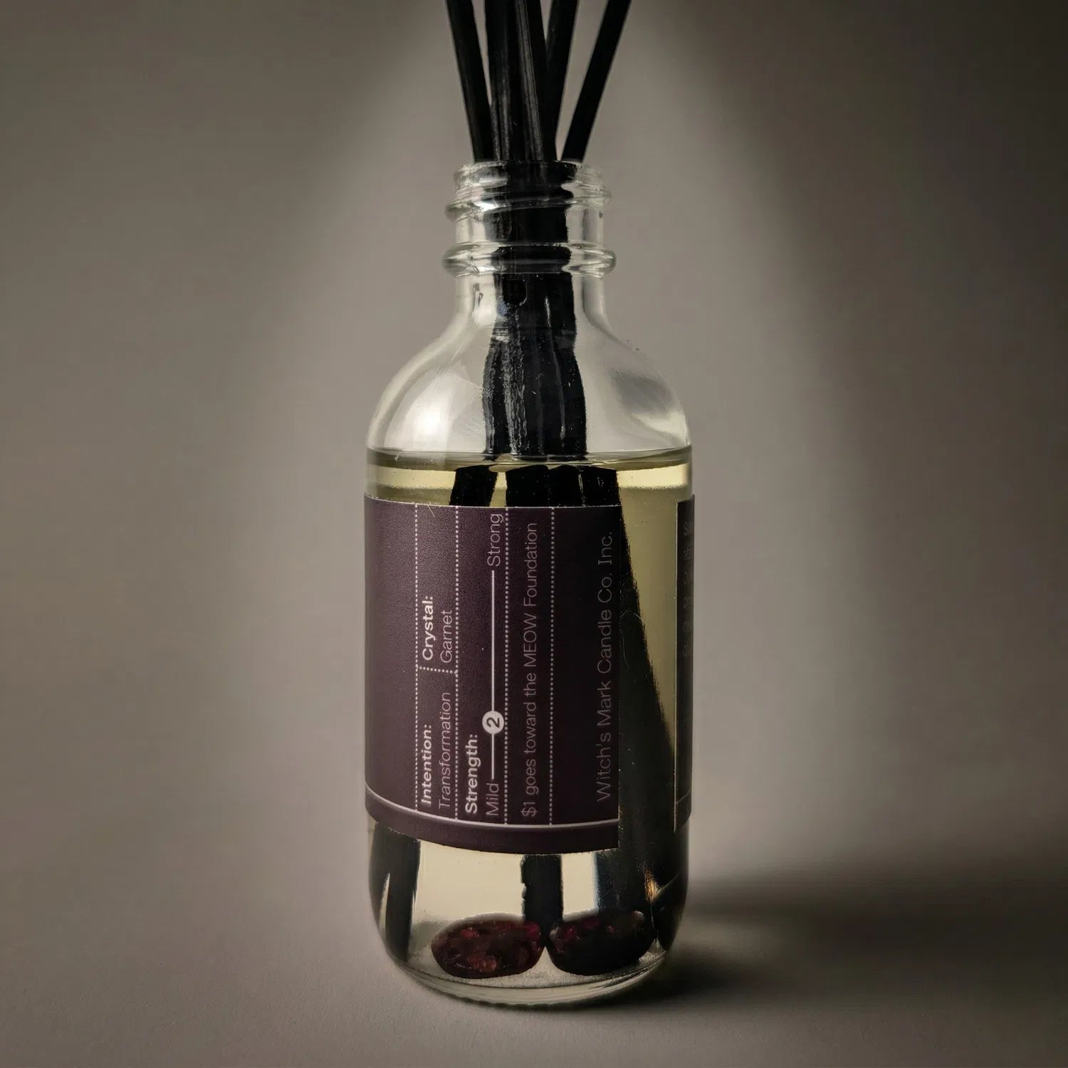 A warmly lit close-up of the Hecate spell jar reed diffuser, revealing the fierce garnet crystals submerged at the bottom of the clear botanical oil, crafted to entice higher thinking, self-empowerment, and metamorphic change.