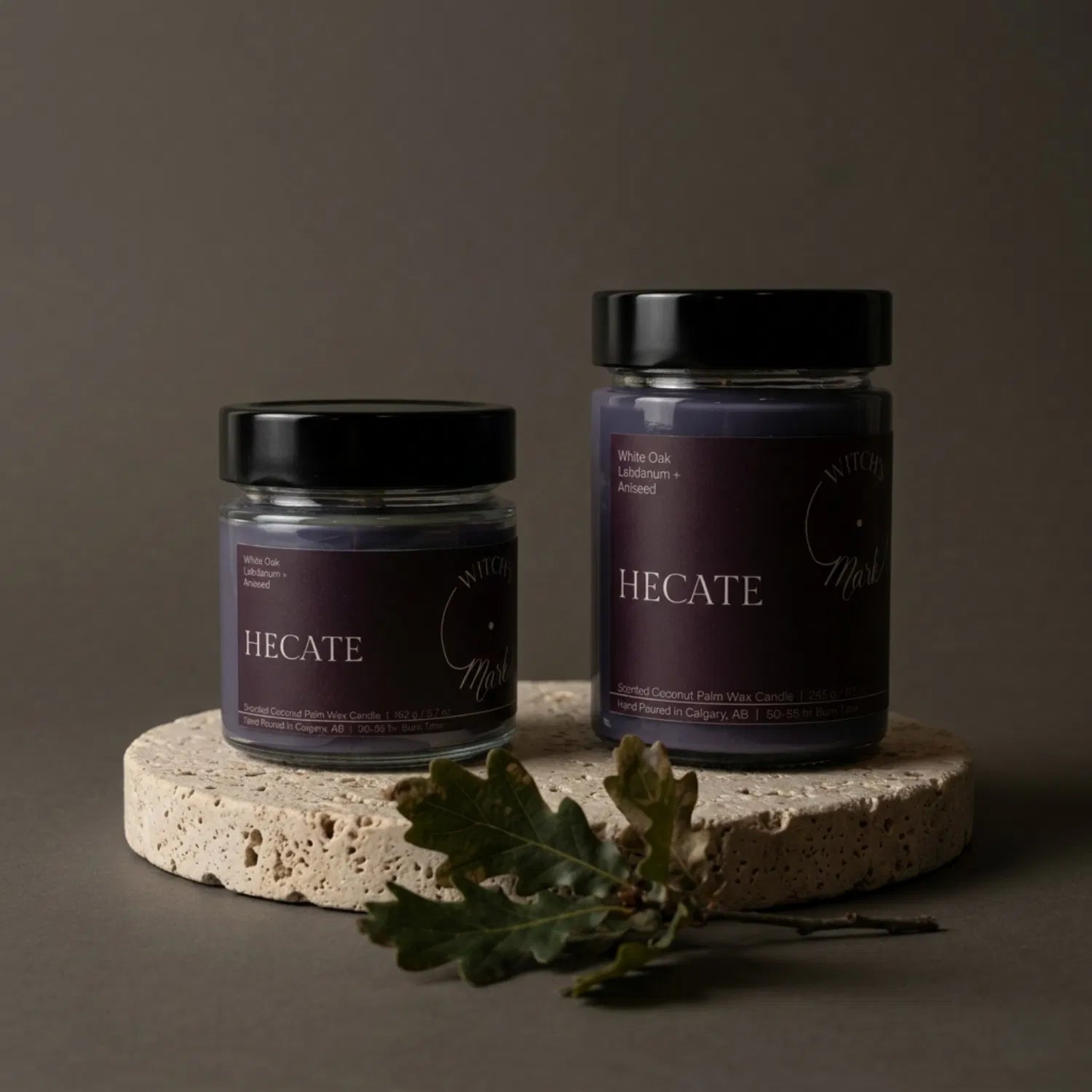 Two sizes of the Witch's Mark Hecate coconut wax candle, 5.7 oz and 8.6 oz, displaying the deep purple wax on a round stone base, garnished with grounding white oak leaves.