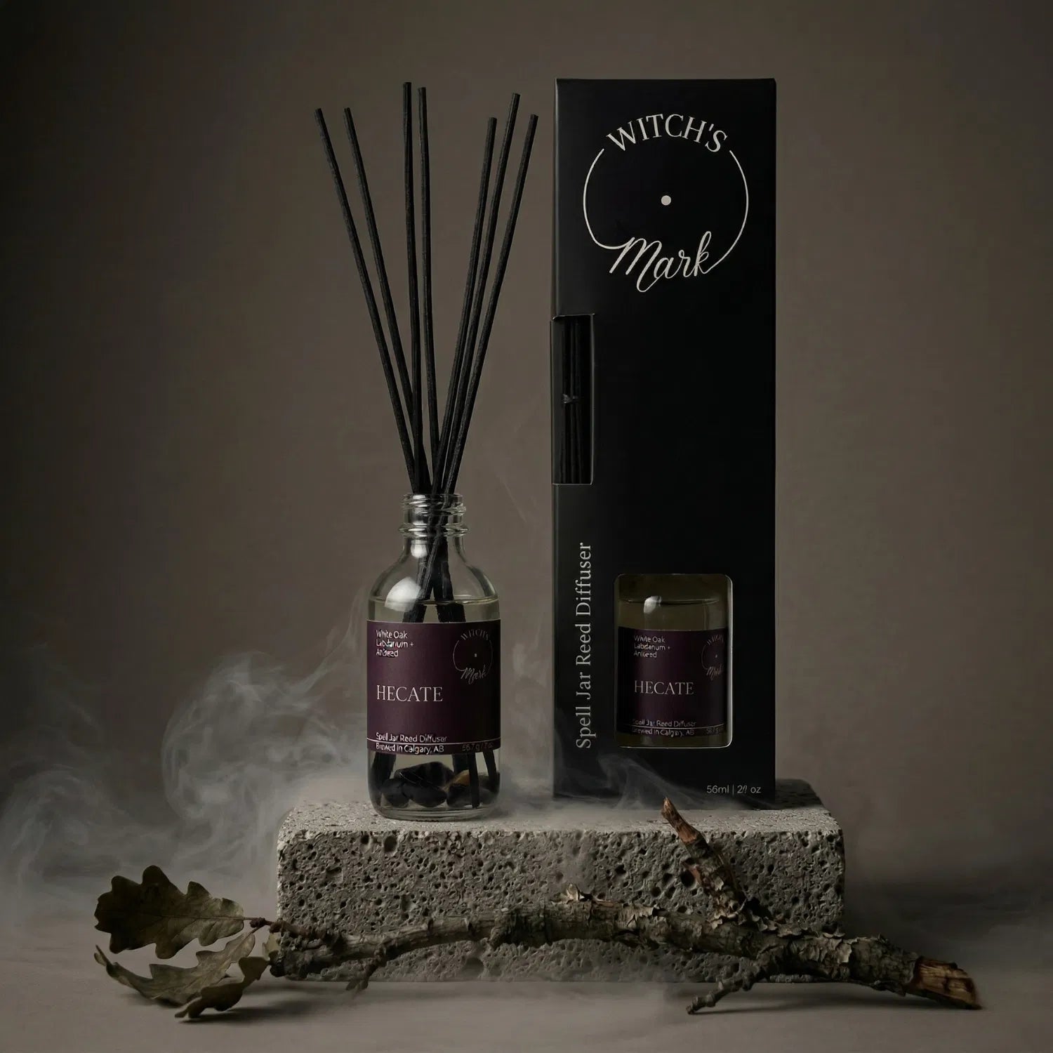 The Hecate spell jar reed diffuser by Witch's Mark displayed next to its sleek black packaging box. The apothecary glass vessel with black reeds rests on a textured stone block surrounded by mystical mist and a grounding white oak branch, crafted to guide you through profound transformation.