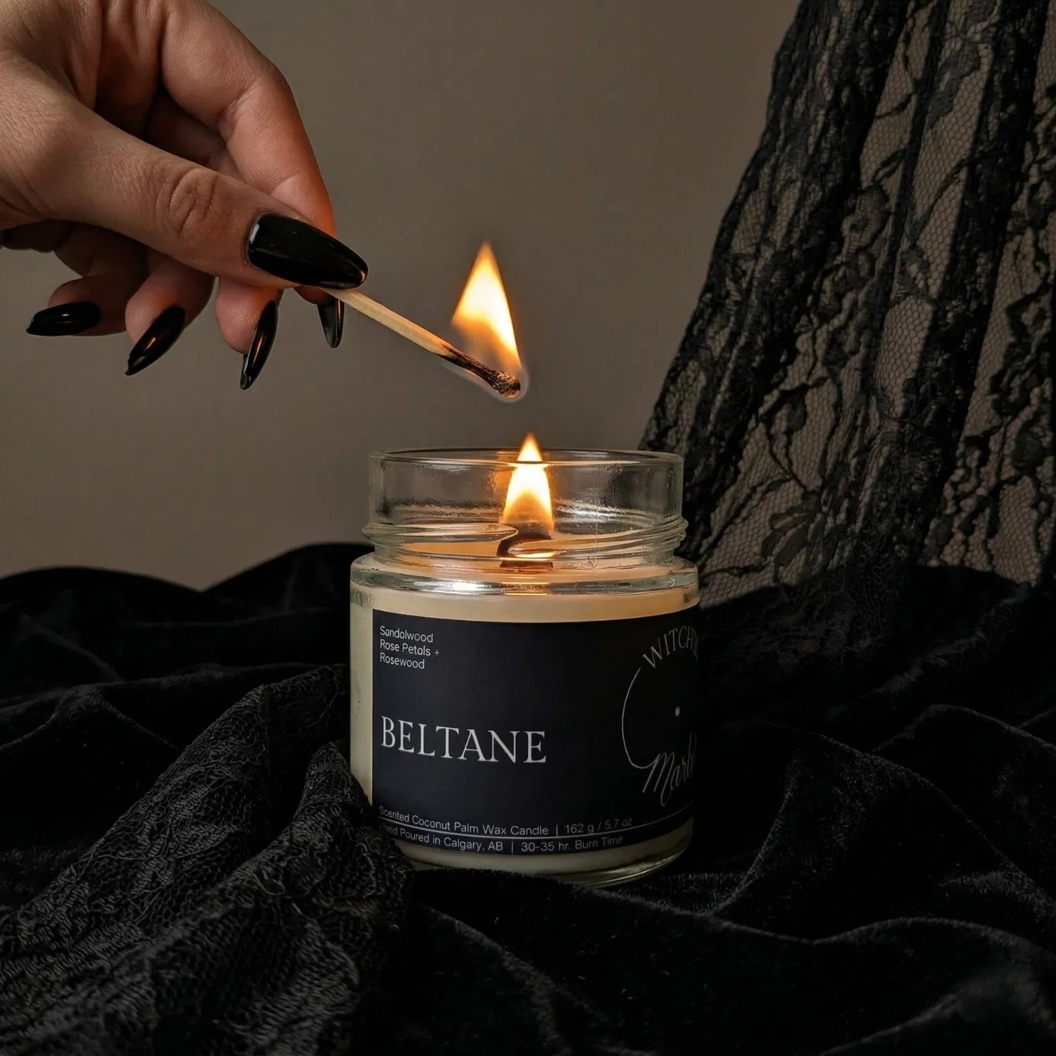 A hand with black nail polish using a match to light the wooden wick of a seasonal Beltane May Day candle by Witch's Mark, set against a dark aesthetic background featuring draped black lace.