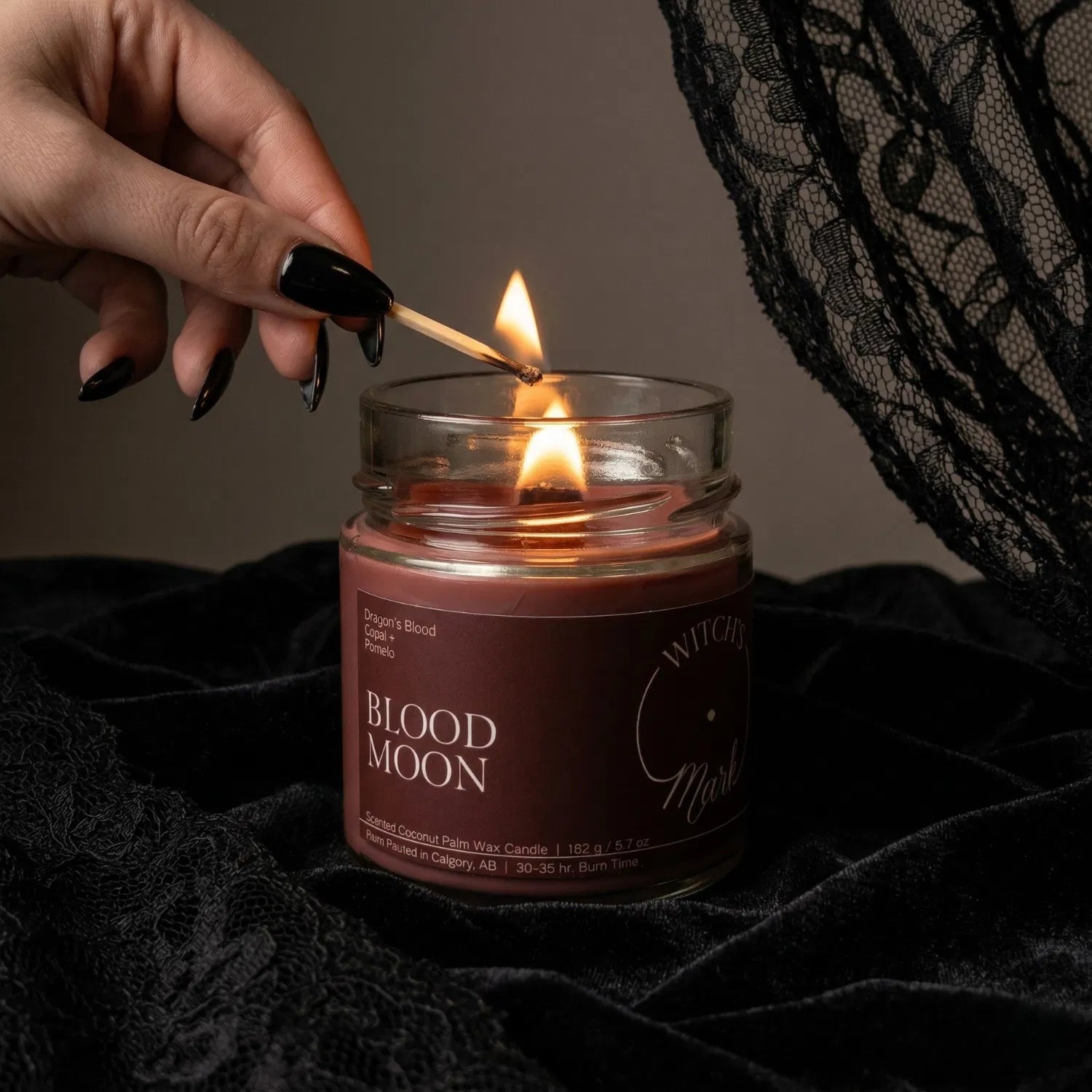 A hand with black nail polish using a match to light the wooden wick of a deep red Blood Moon protection candle by Witch's Mark, set against a dark aesthetic background featuring draped black lace.
