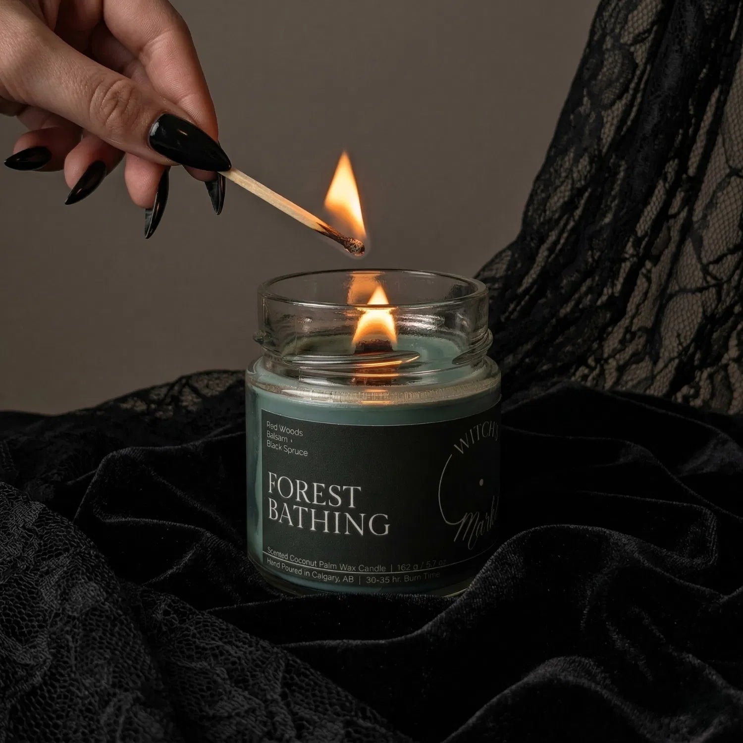 A hand with black nail polish using a match to light the wooden wick of a deep green Forest Bathing candle by Witch's Mark, set against a dark aesthetic background featuring draped black lace.