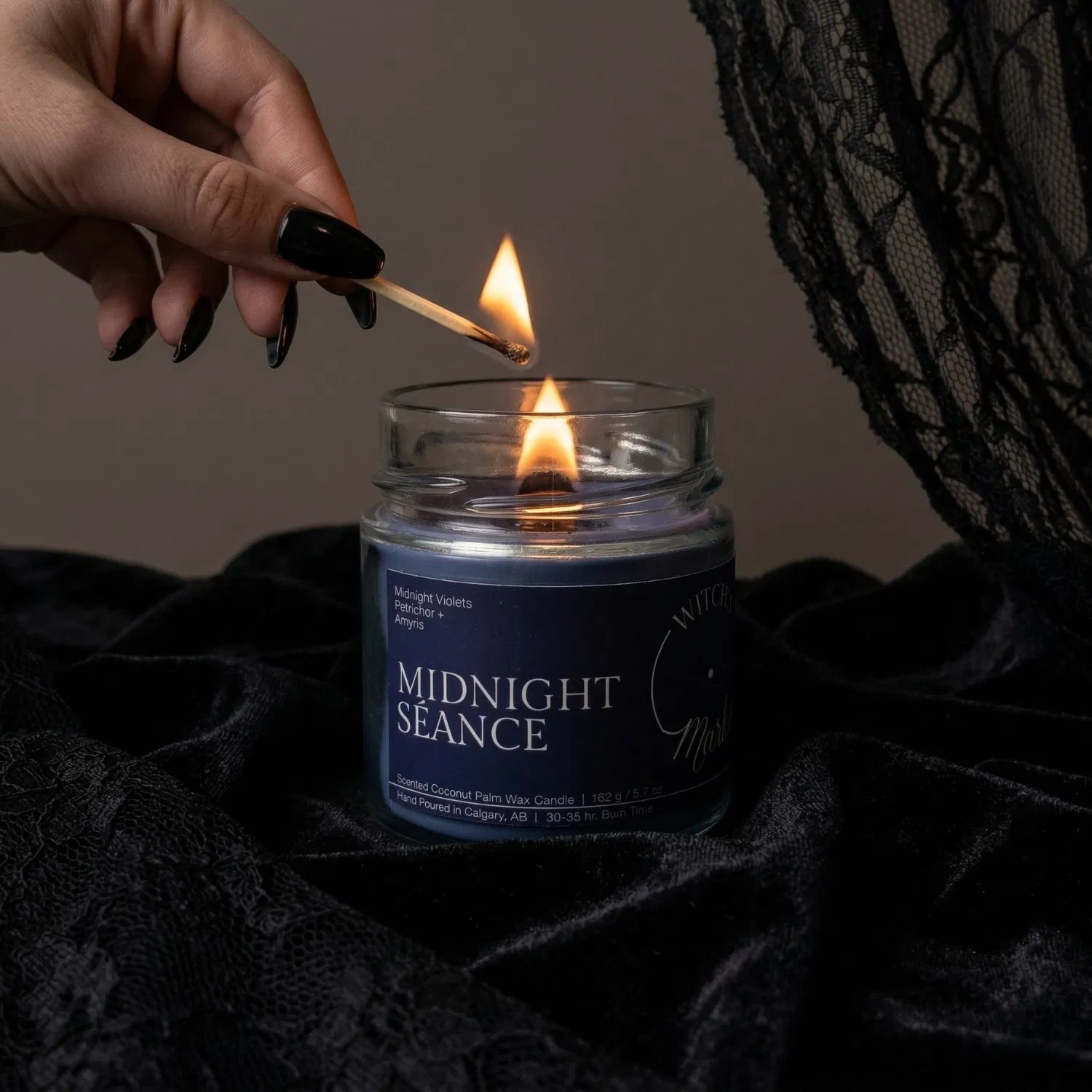 A hand with black nail polish using a match to light the wooden wick of a blue Midnight Séance spiritual candle by Witch's Mark, set against a dark aesthetic background featuring draped black lace.