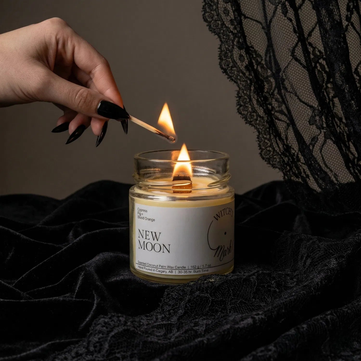 A hand with black nail polish using a match to light the wooden wick of a New Moon intention candle by Witch's Mark, set against a dark aesthetic background featuring draped black lace.