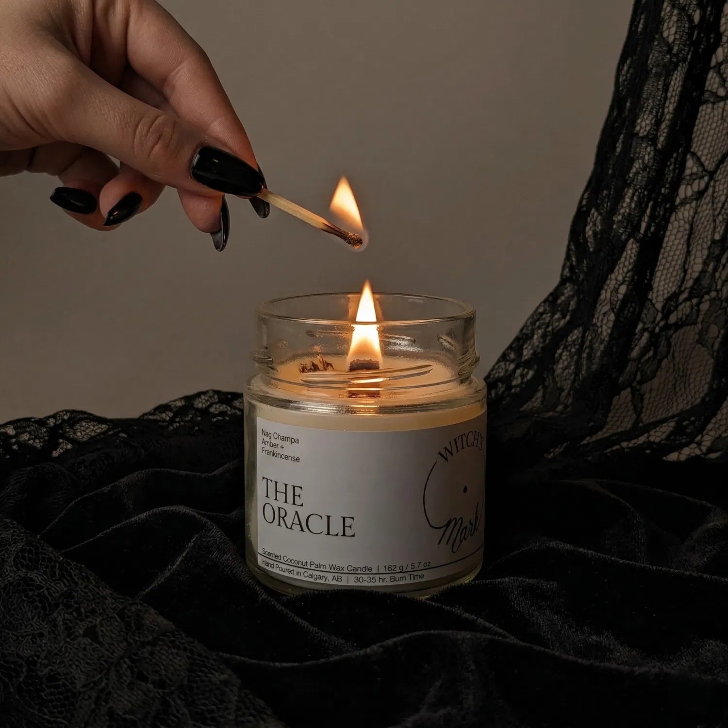 A hand with black nail polish using a match to light the wooden wick of an Oracle tarot candle by Witch's Mark, set against a dark aesthetic background featuring draped black lace.
