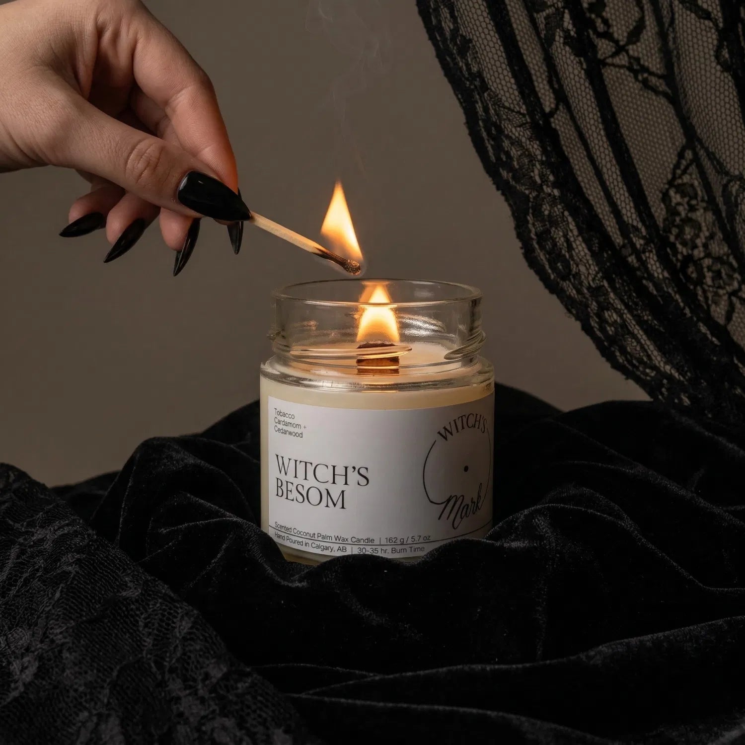 A hand with black nail polish using a match to light the wooden wick of a Witch's Besom protection candle by Witch's Mark, set against a dark aesthetic background featuring draped black lace.