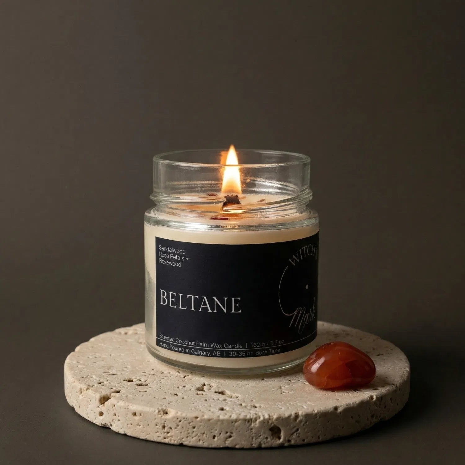 A lit Beltane crystal candle by Witch's Mark resting on a textured stone coaster next to a vibrant orange carnelian crystal, crafted to inspire passion, creativity, and confidence.