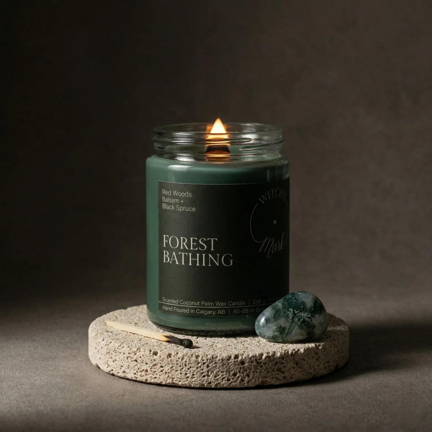 A lit Forest Bathing crystal candle with deep green wax from the Rooted Intentions Bundle, set on a stone coaster alongside a grounding tree agate crystal to help anchor energy and stay rooted in purpose.