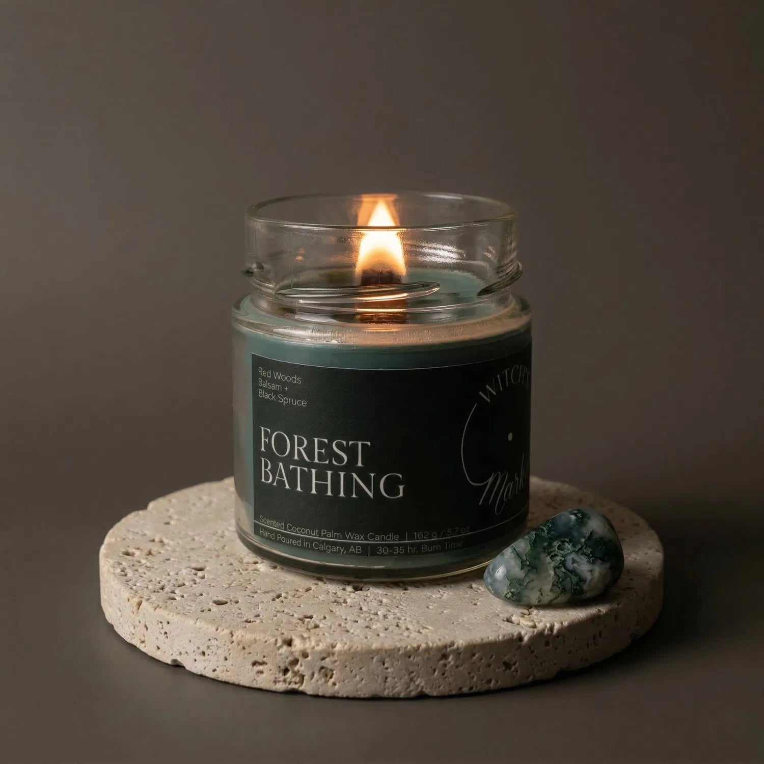 A lit Forest Bathing crystal candle by Witch's Mark resting on a textured stone coaster next to a grounding tree agate crystal. The deep green wax is designed to promote balance and a deep connection to nature.