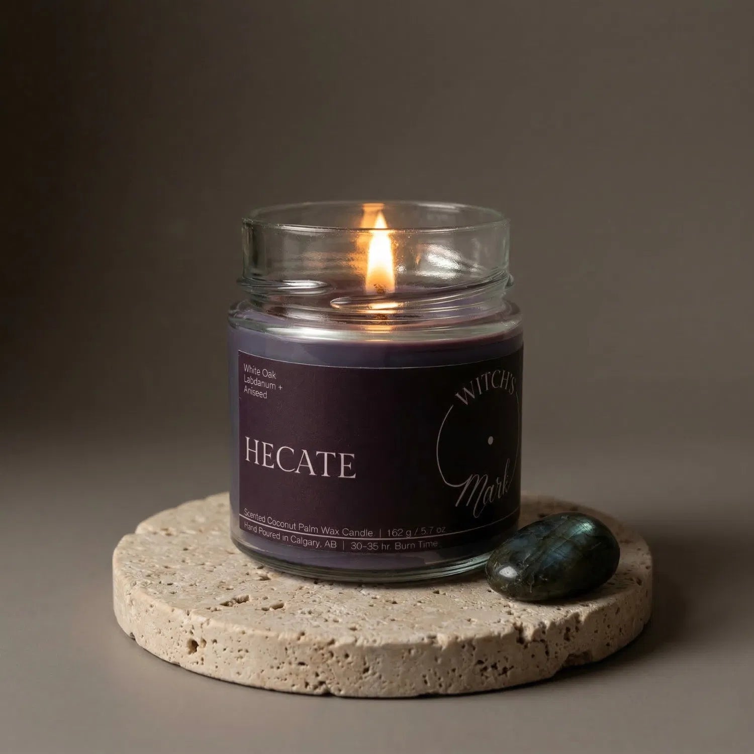 A lit Hecate crystal candle by Witch's Mark resting on a textured stone coaster next to a labradorite crystal. The deep purple wax is designed to promote transformation and personal growth during magical rituals.