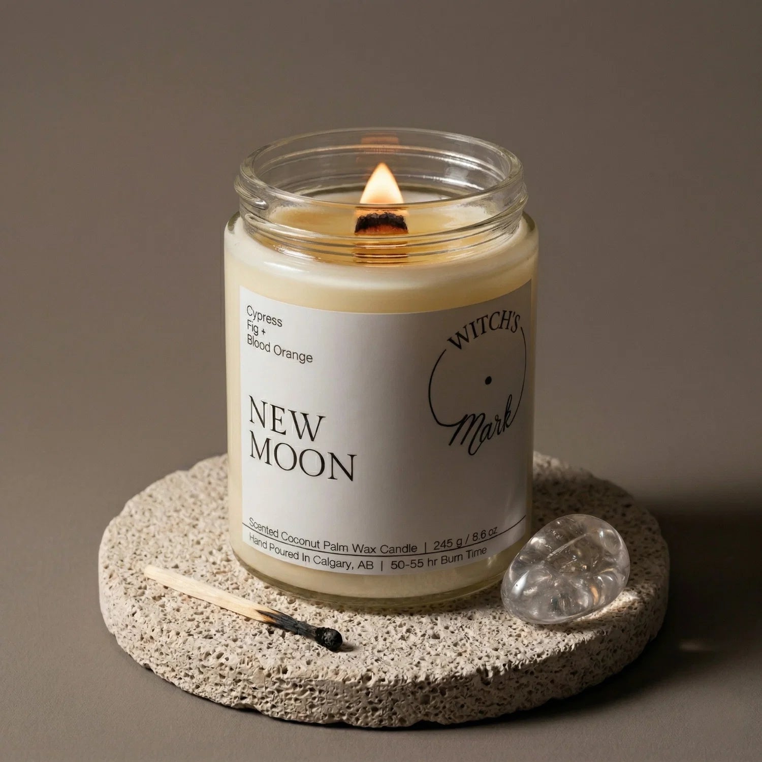 A lit New Moon crystal candle from the Rooted Intentions Bundle, resting on a stone coaster next to a clear quartz crystal used for sparking clarity and focusing goals.