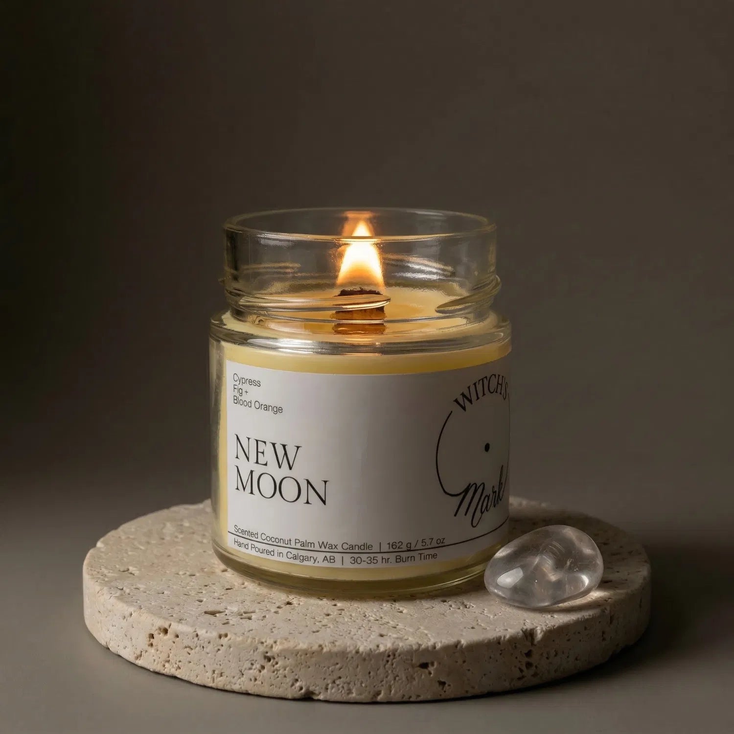 A lit New Moon crystal candle by Witch's Mark resting on a textured stone coaster next to a smooth clear quartz crystal, designed to help focus and set intentions.