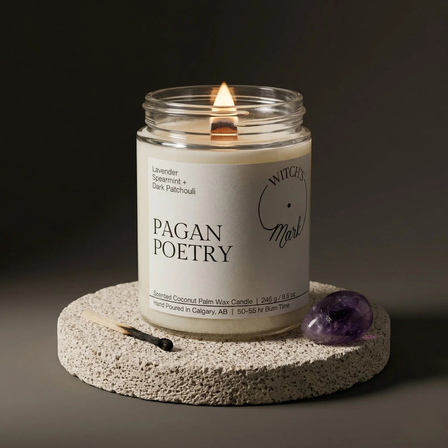 A lit Pagan Poetry crystal candle from the Release and Restore Bundle, set on a stone coaster alongside a purple amethyst crystal for deep grounding and energy restoration.