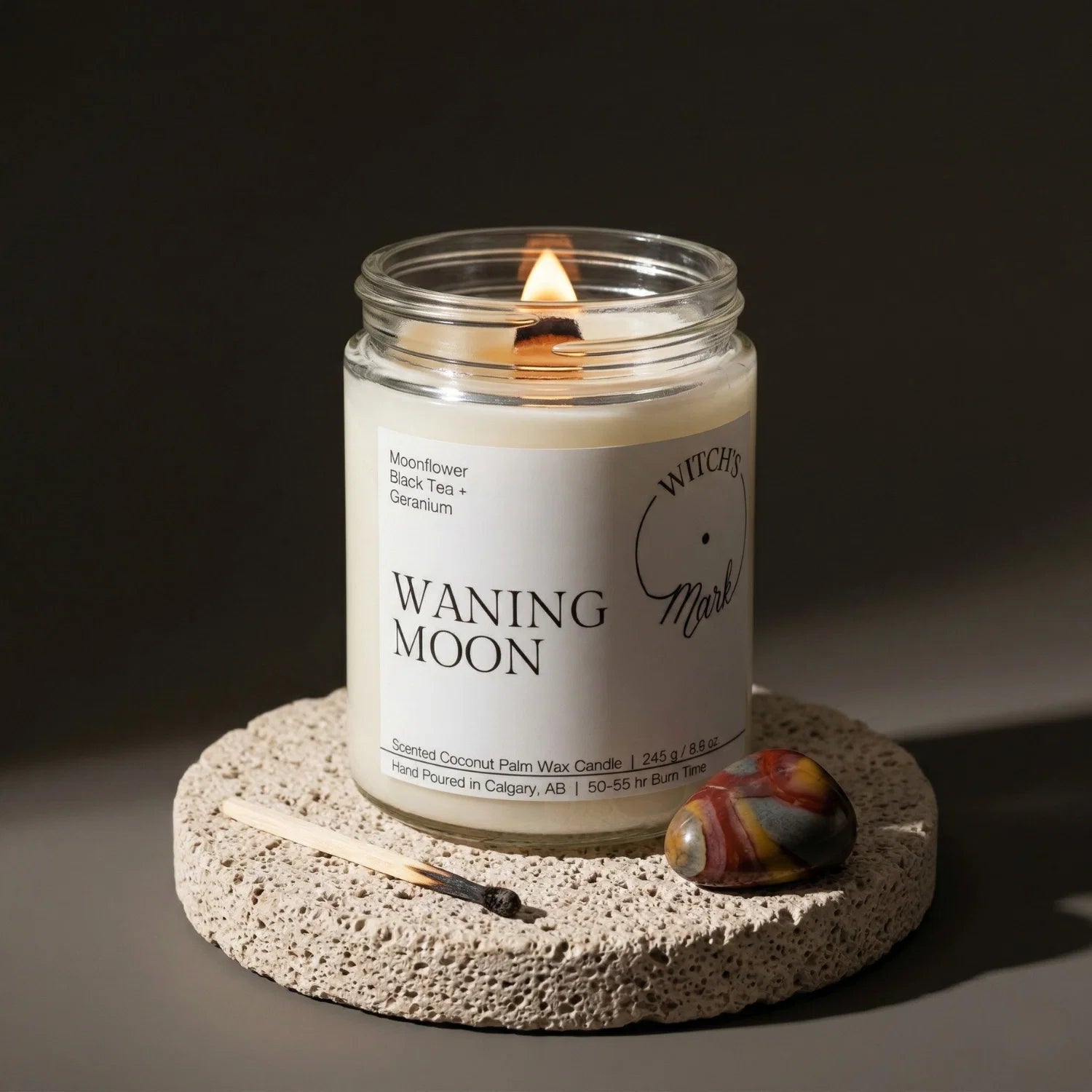 A lit Waning Moon candle from the Lunar Cycle bundle resting on a stone coaster, alongside a vibrant polychrome jasper crystal crafted to aid in letting go and cleansing old habits.