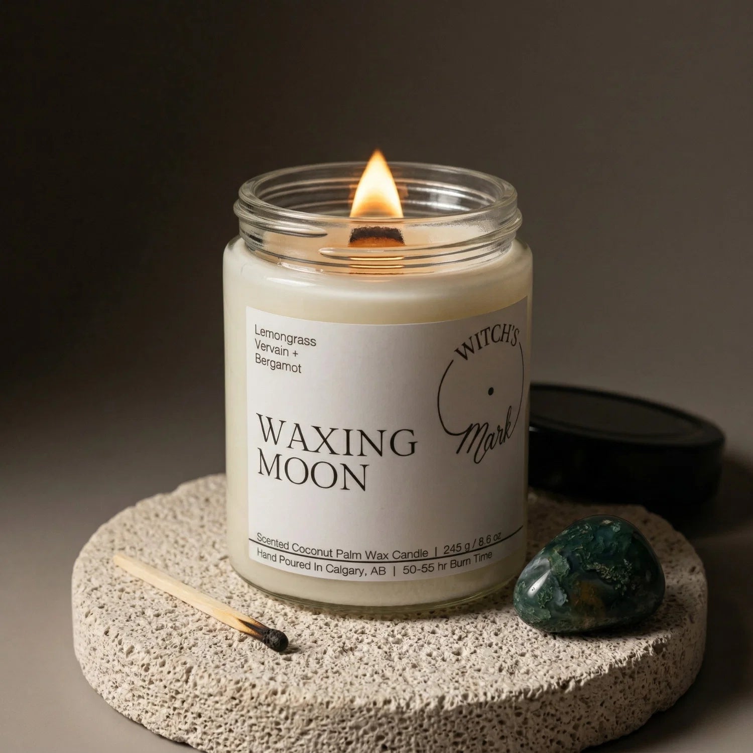 A lit Waxing Moon candle from the Lunar Cycle bundle resting on a stone coaster, next to a green moss agate crystal used for building momentum and attracting abundance.