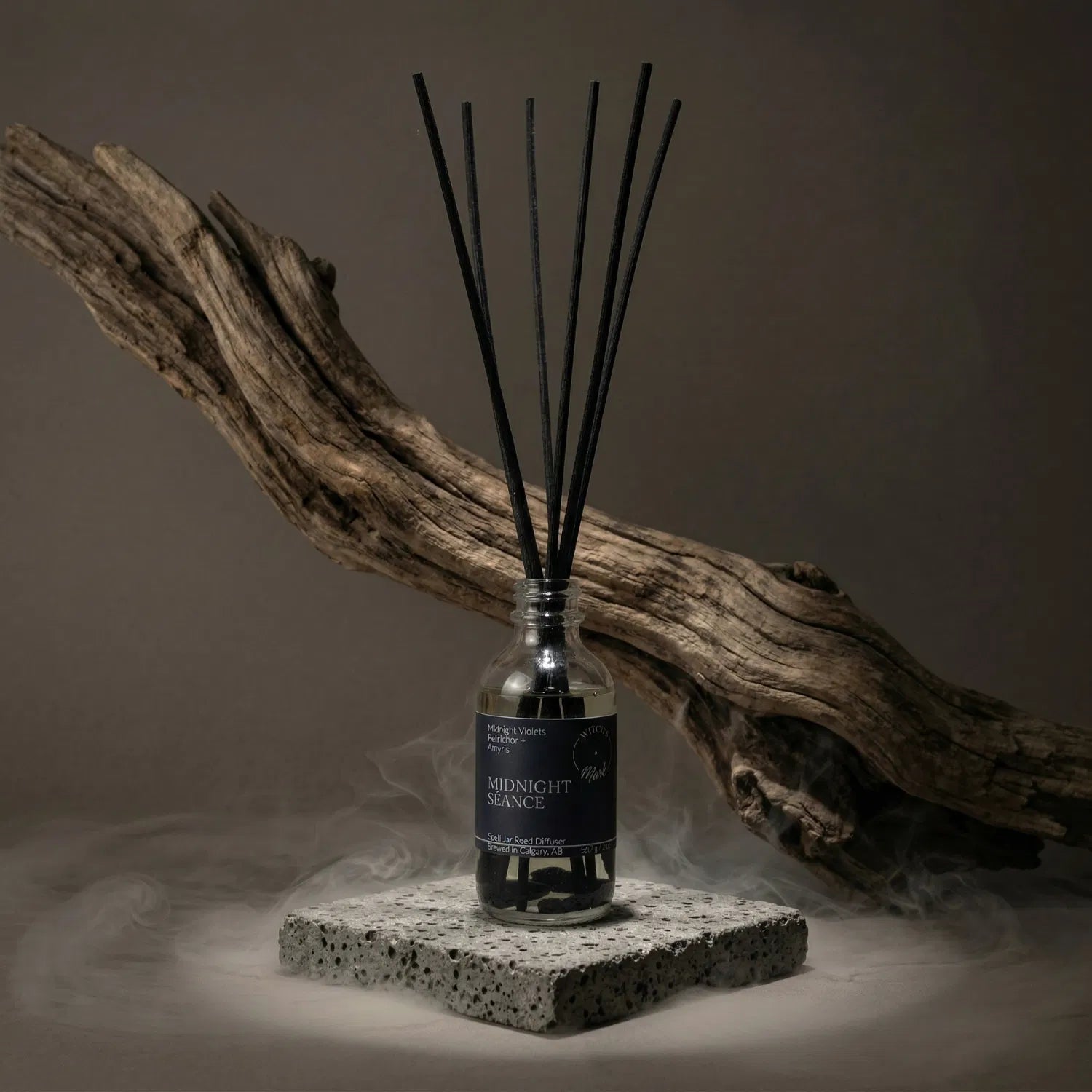 The Midnight Séance apothecary spell jar reed diffuser by Witch's Mark sitting on a stone base, enveloped in atmospheric mist with natural driftwood in the background, perfectly setting the stage for deep divination and astral travel.