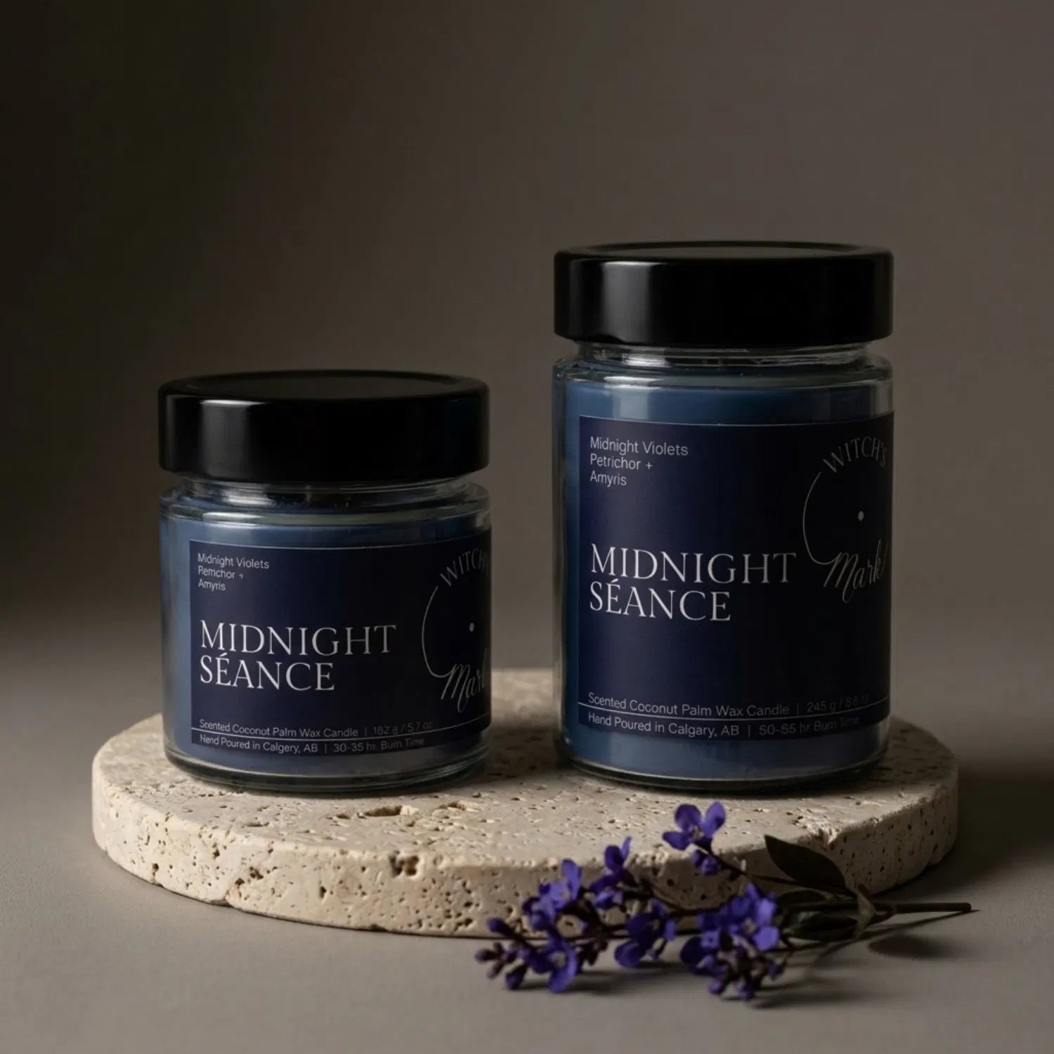 Two sizes of the Witch's Mark Midnight Séance coconut wax candle, 5.7 oz and 8.6 oz, displaying the deep blue, glitter-adorned wax on a round stone base, garnished with fresh midnight violet flowers.