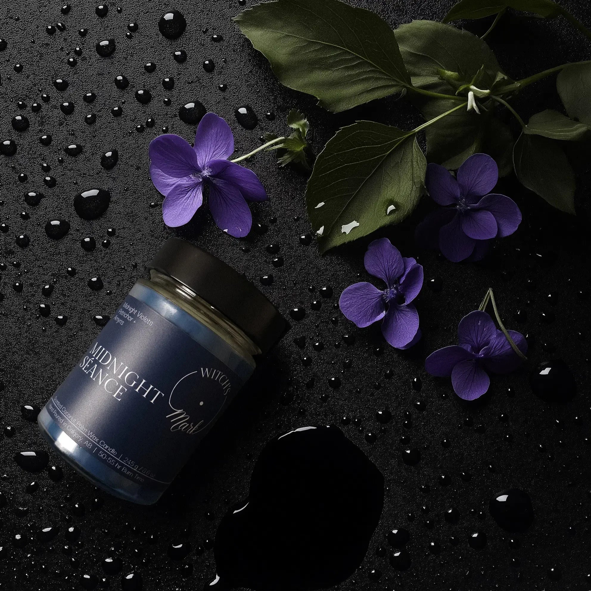 A top-down flat lay highlighting the mystical scent profile of the Midnight Séance candle, featuring the blue wax jar surrounded by fresh midnight violets and water droplets representing earthy petrichor on a dark slate background.