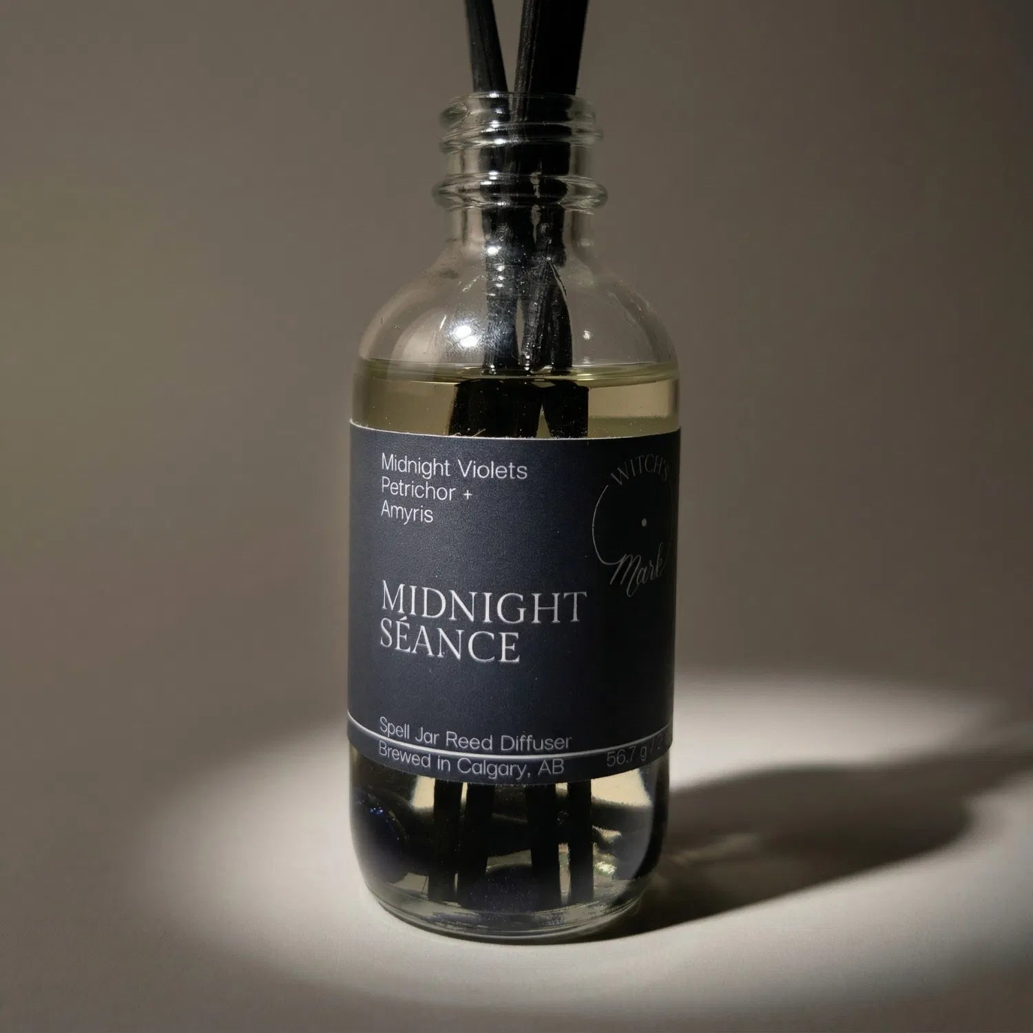 A dramatic spotlight close-up of the Midnight Séance spell jar reed diffuser, highlighting the dark blue front label which lists its magical, illuminating scent profile of haunting midnight violets, earthy petrichor, and resinous amyris.