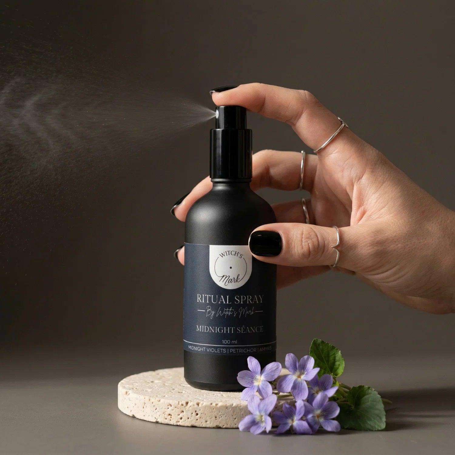 A hand with black nail polish spritzing the Witch's Mark Midnight Séance ritual spray over fresh purple midnight violets. This dark apothecary aura mist is infused with blue goldstone charged moon water to commune with the unseen and empower nocturnal spirit work.
