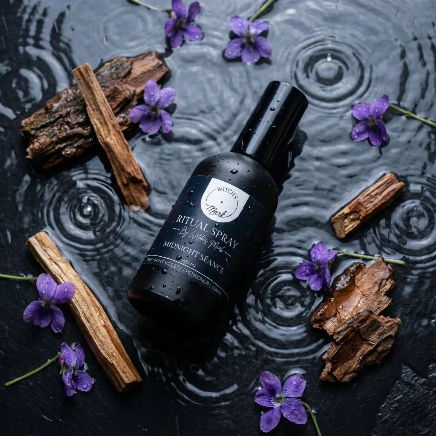 A top-down flat lay highlighting the otherworldly ingredients of the Midnight Séance ritual mist by Witch's Mark. The black apothecary bottle sits on a wet, rippling dark slate surface representing earthy petrichor, surrounded by fresh midnight violets and pieces of resinous amyris wood.