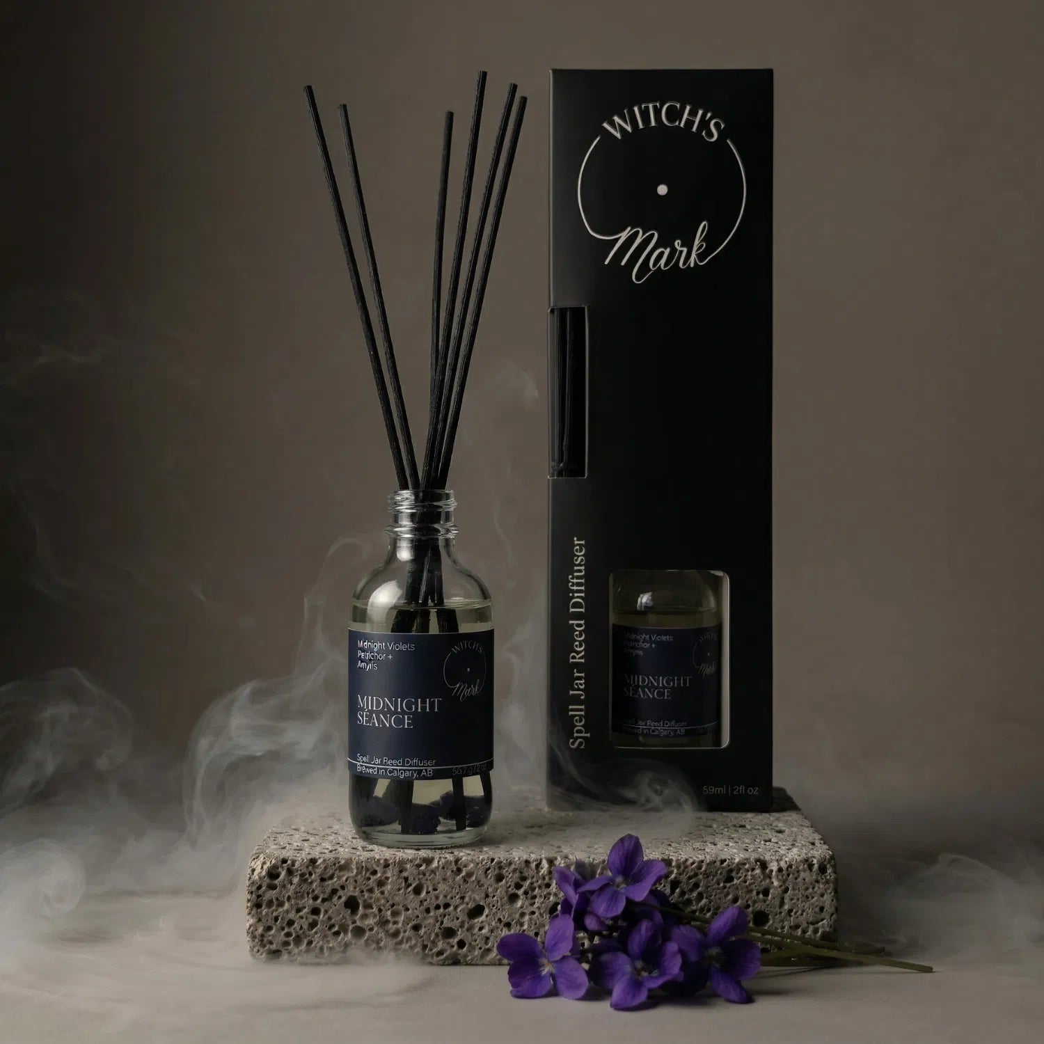 The Midnight Séance spell jar reed diffuser by Witch's Mark displayed next to its sleek black packaging box. The apothecary glass vessel with black reeds rests on a textured stone block surrounded by mystical mist and fresh purple midnight violets, crafted to commune with the unseen.