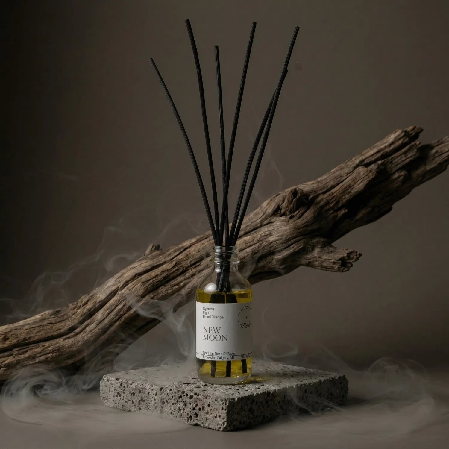 A New Moon apothecary spell jar reed diffuser by Witch's Mark sitting on a stone base, surrounded by atmospheric mist and natural driftwood, perfectly capturing the fertile darkness of the lunar cycle for intention setting.